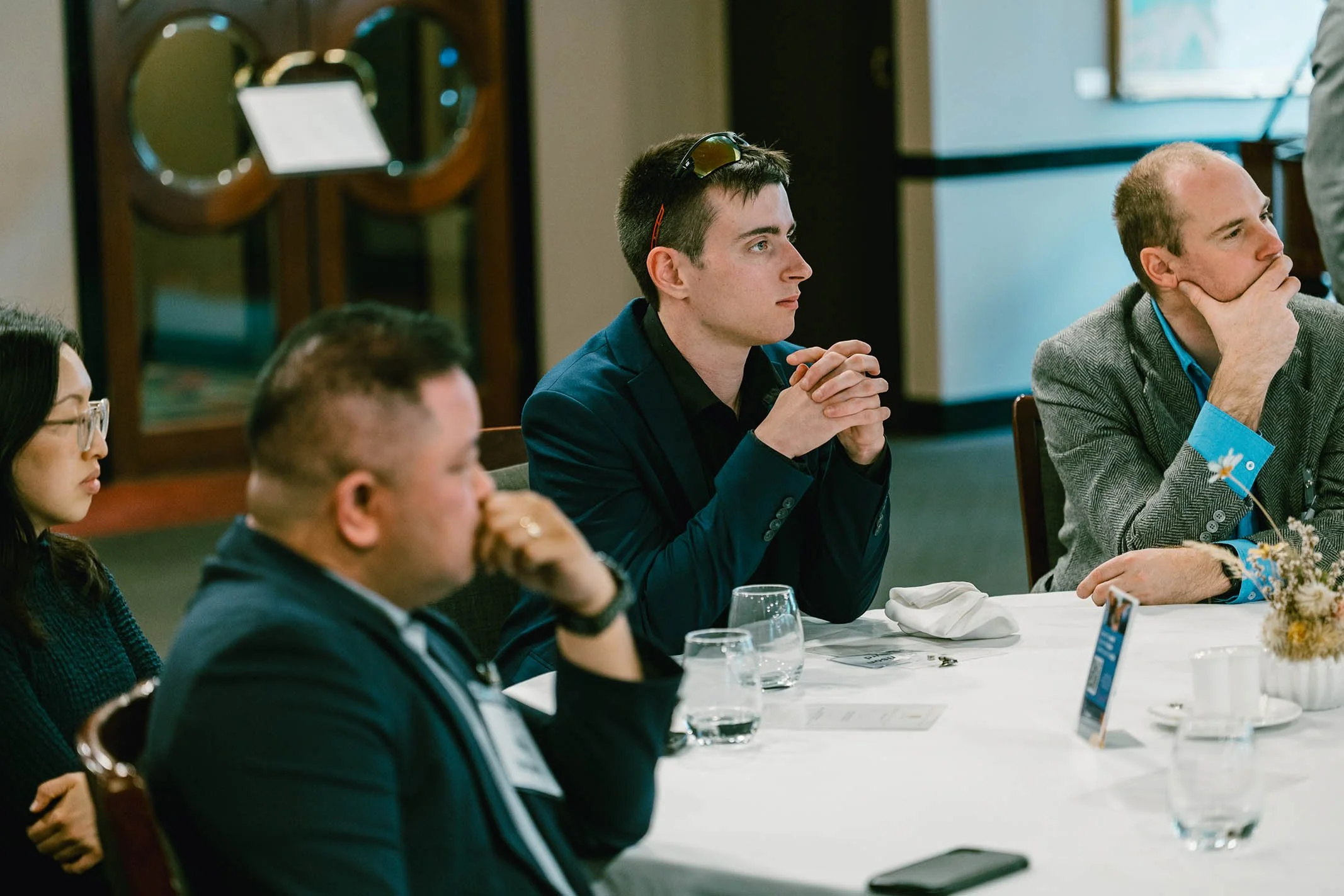 People sitting at a conference table, listening attentively. The central figure is a young man with short dark hair wearing a dark blazer, with sunglasses resting on his head. To his right is a man with a wristband and a flower on his blazer, resting