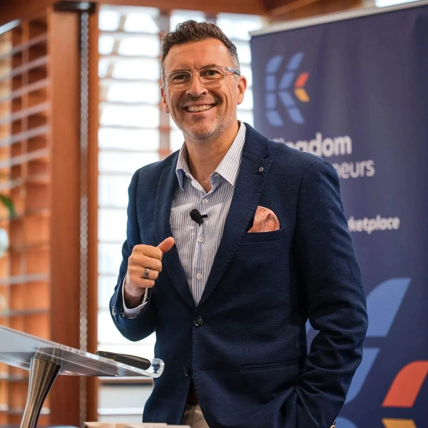 Confident man in blazer smiling at camera during presentation with a podium and banner in the background.