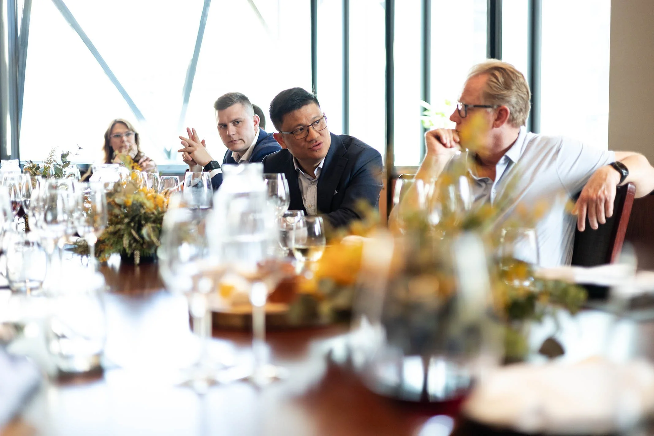 People sitting at a long table during a meeting or conference, with glasses and plates on the table, and a large window in the background.