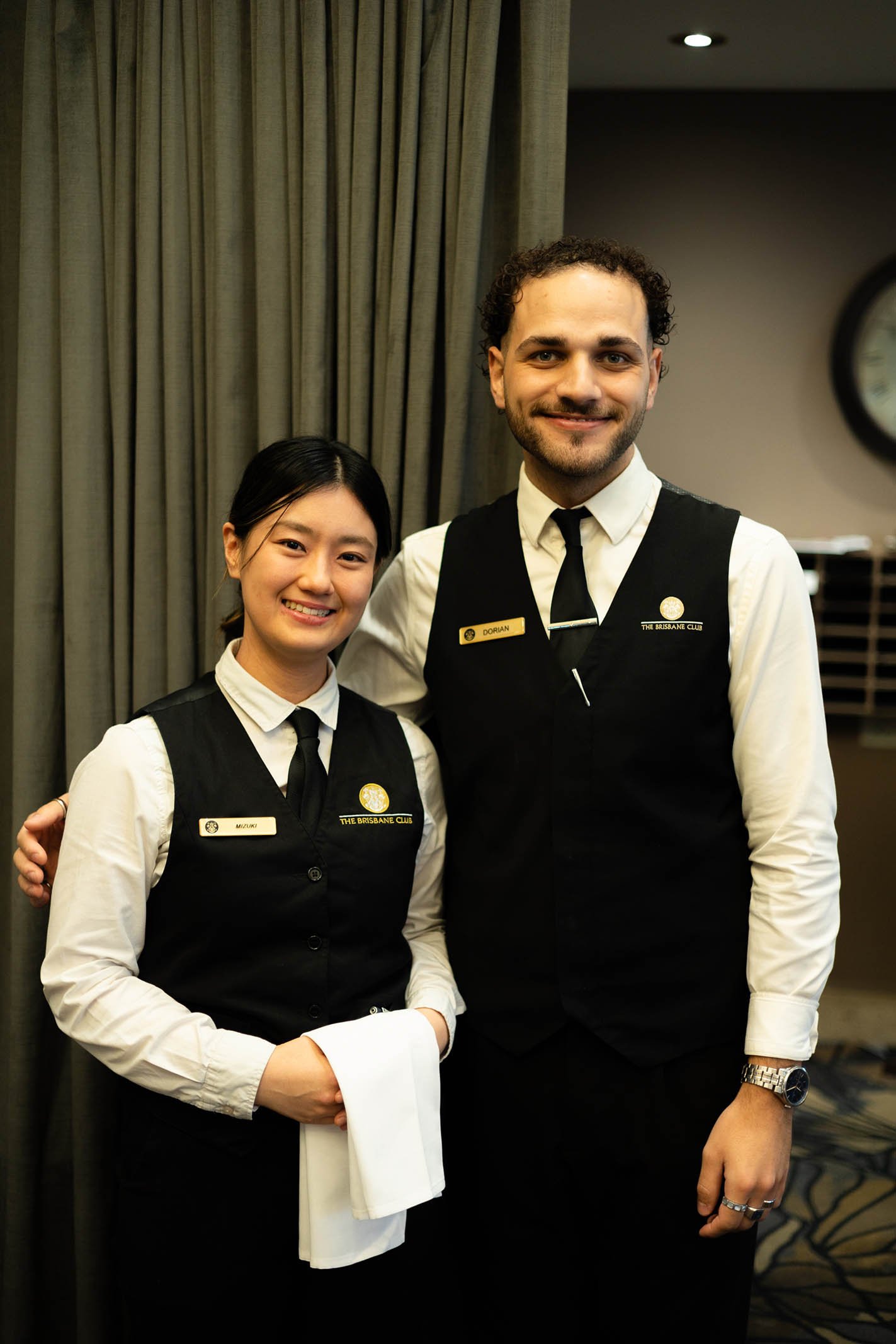 Two restaurant staff members, a woman and a man, in black vests with name tags and white shirts, standing indoors, smiling for the camera.