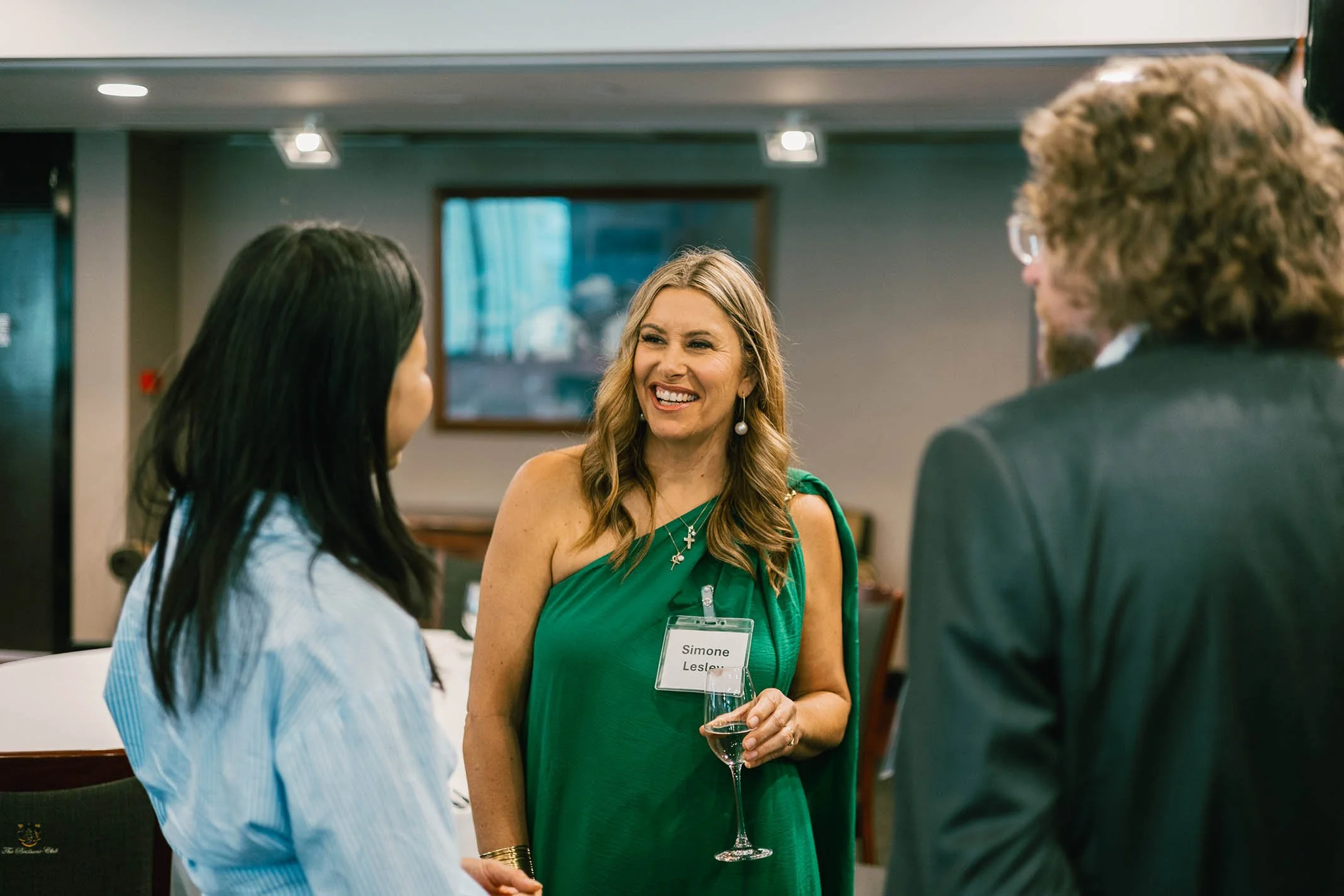 Woman in green dress smiling and holding a glass while talking to two other people in a dimly lit room.