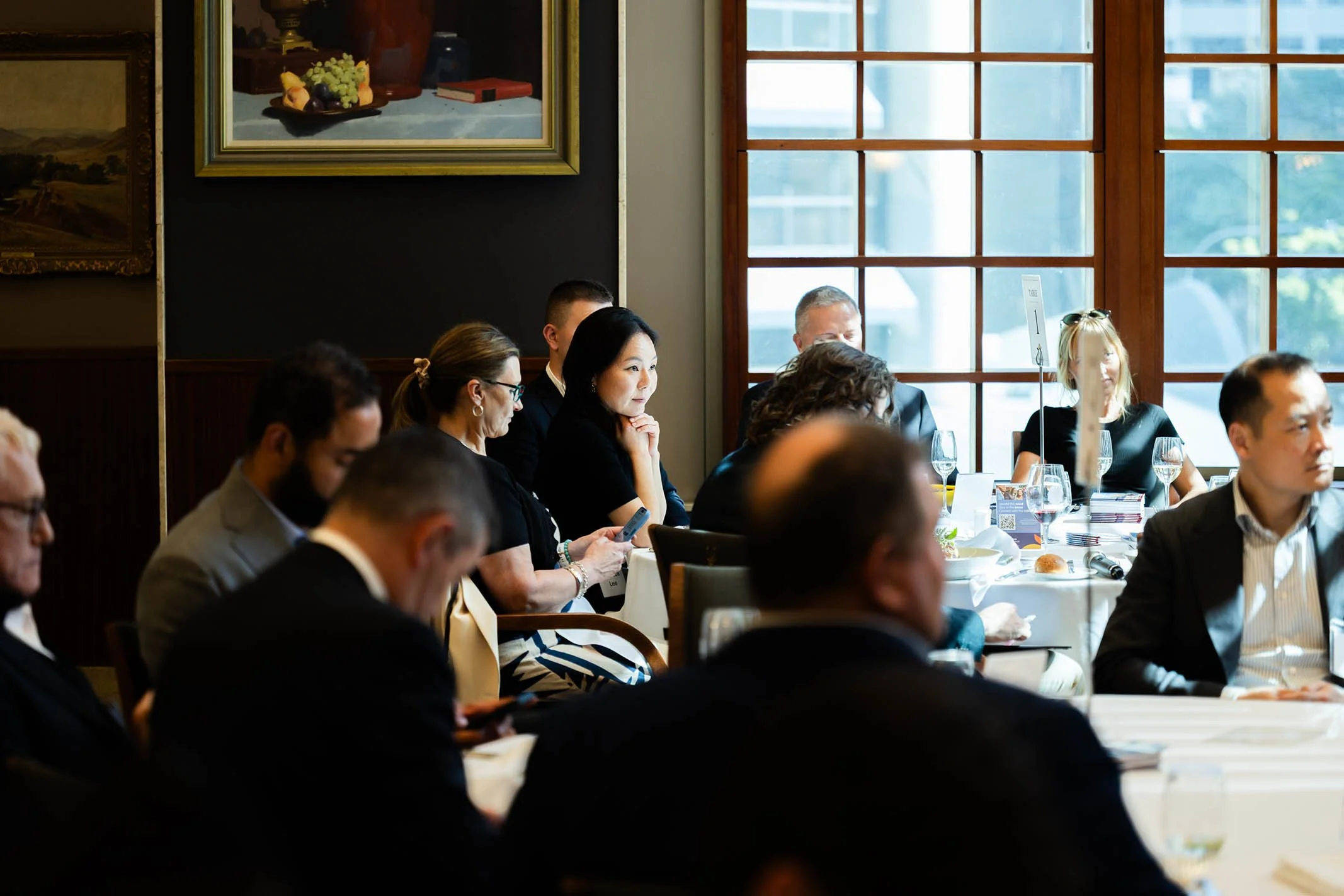 Group of diverse people attending a formal meeting or conference in a well-lit room with large windows, sitting around tables with food and drinks.