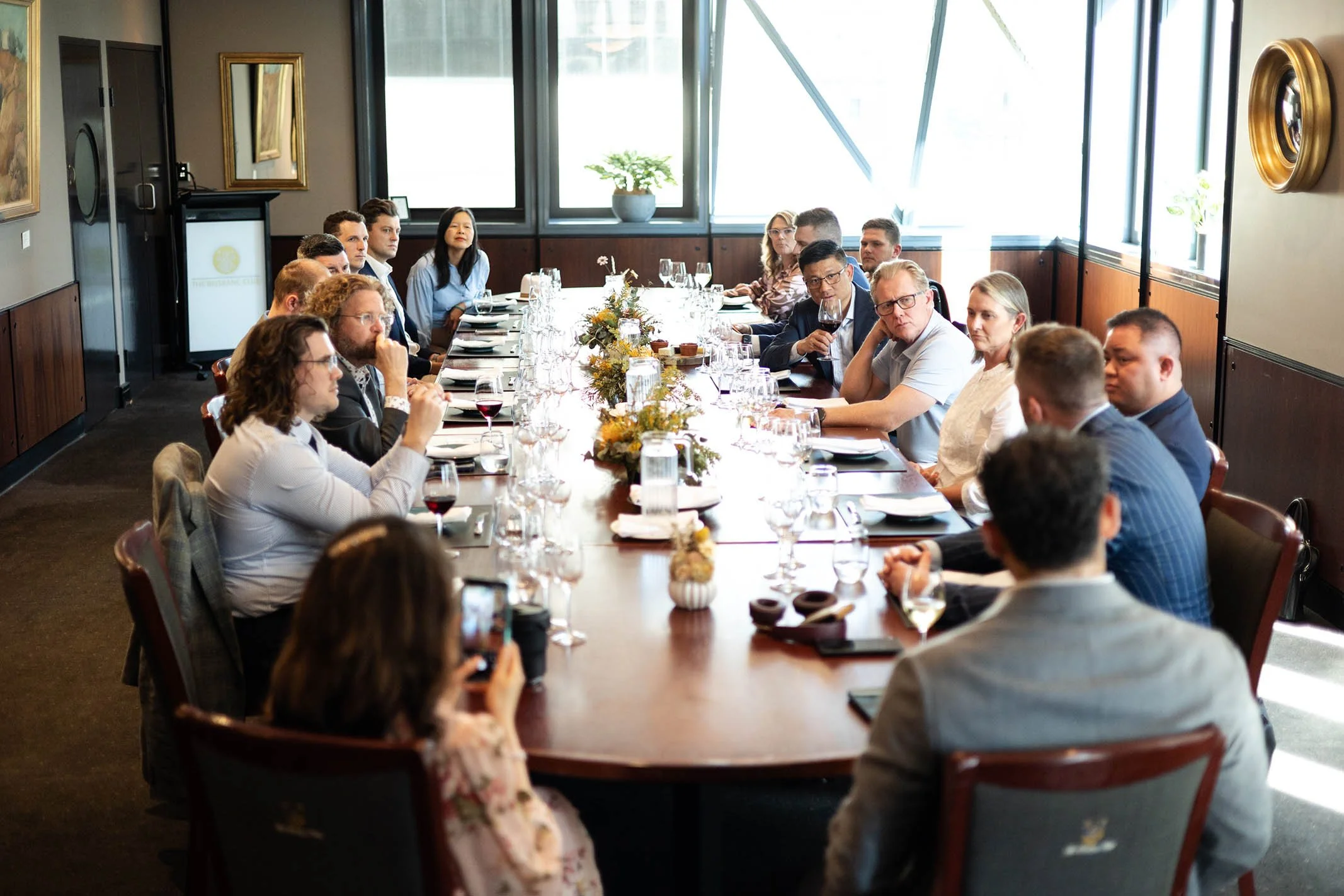 People sitting around a long conference table in a well-lit room with large windows, engaged in a meeting or discussion.