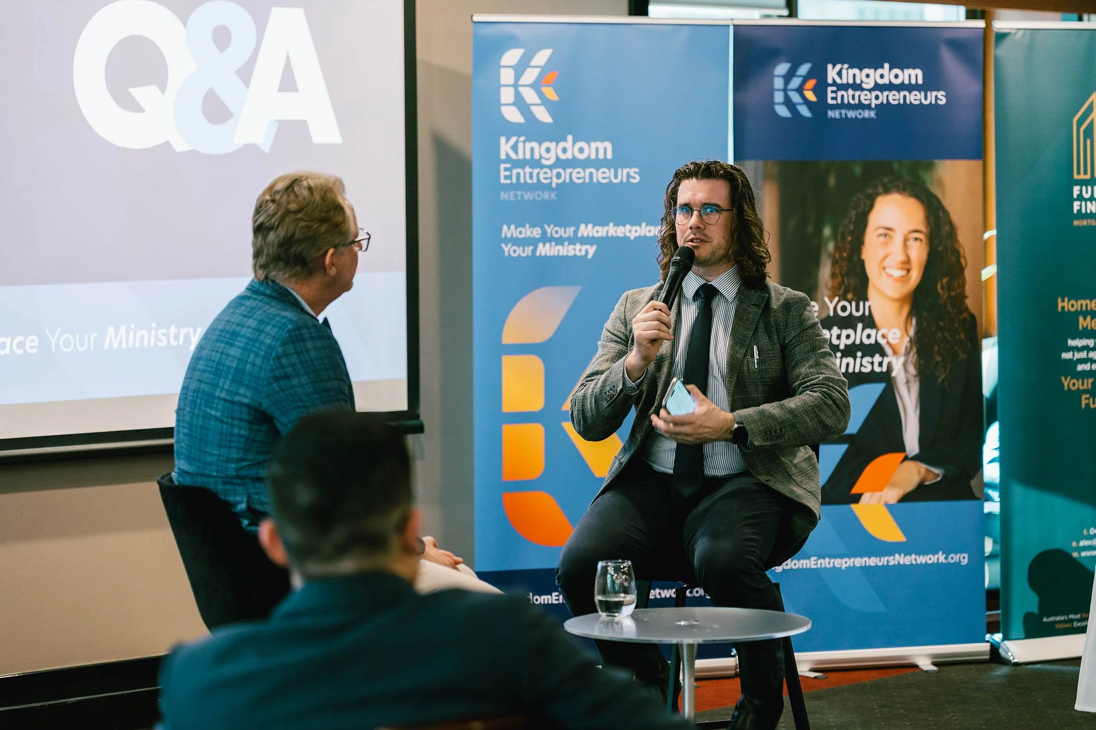 A man with glasses, long hair, and a gray blazer speaking into a microphone during a panel discussion at a Kingdom Entrepreneurs event. An audience is listening, and banners with the event branding are visible in the background.