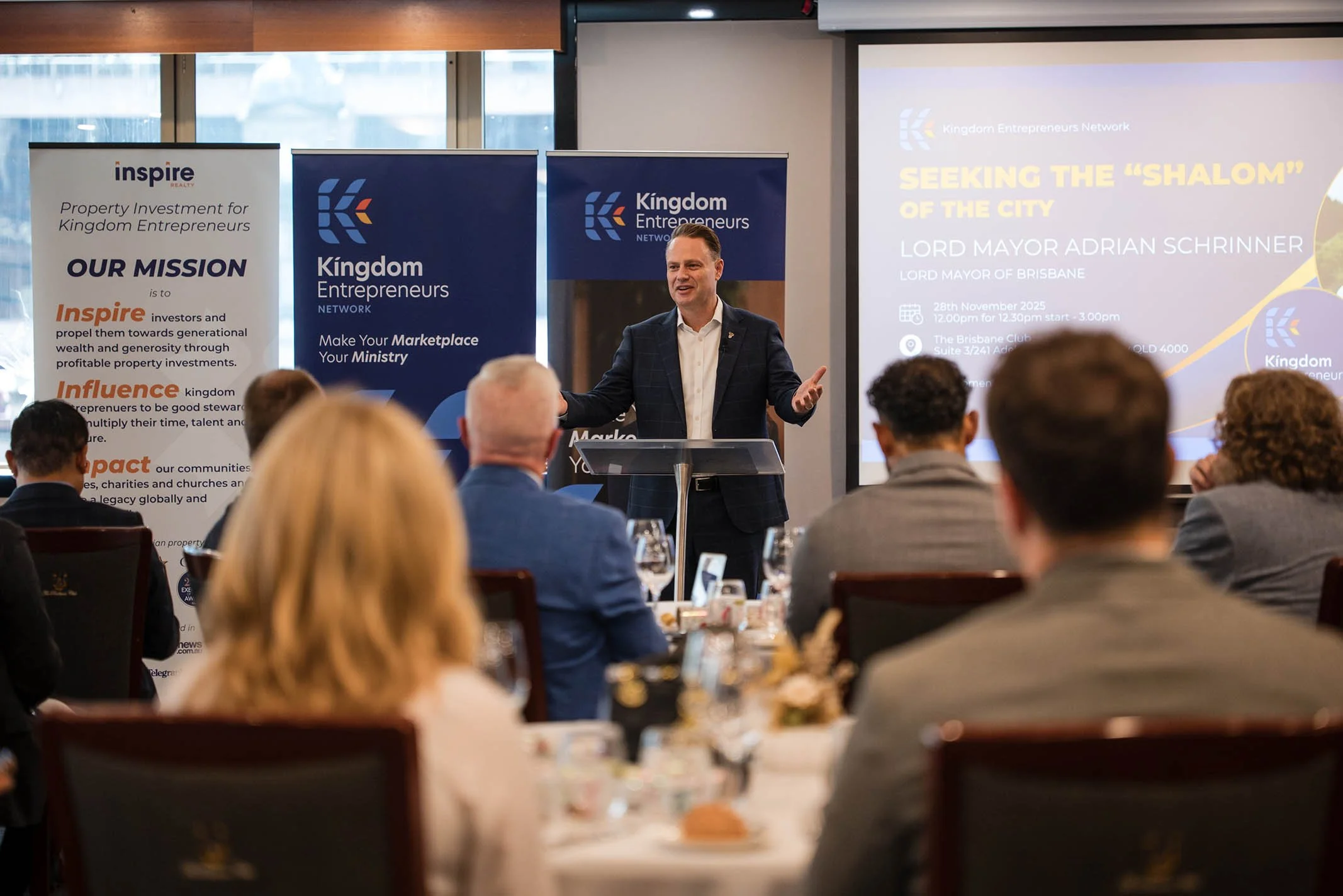 Man giving a presentation at a conference, with a large screen behind him displaying event details and banners on either side with the 'Kingdom Entrepreneurs Network' logo.