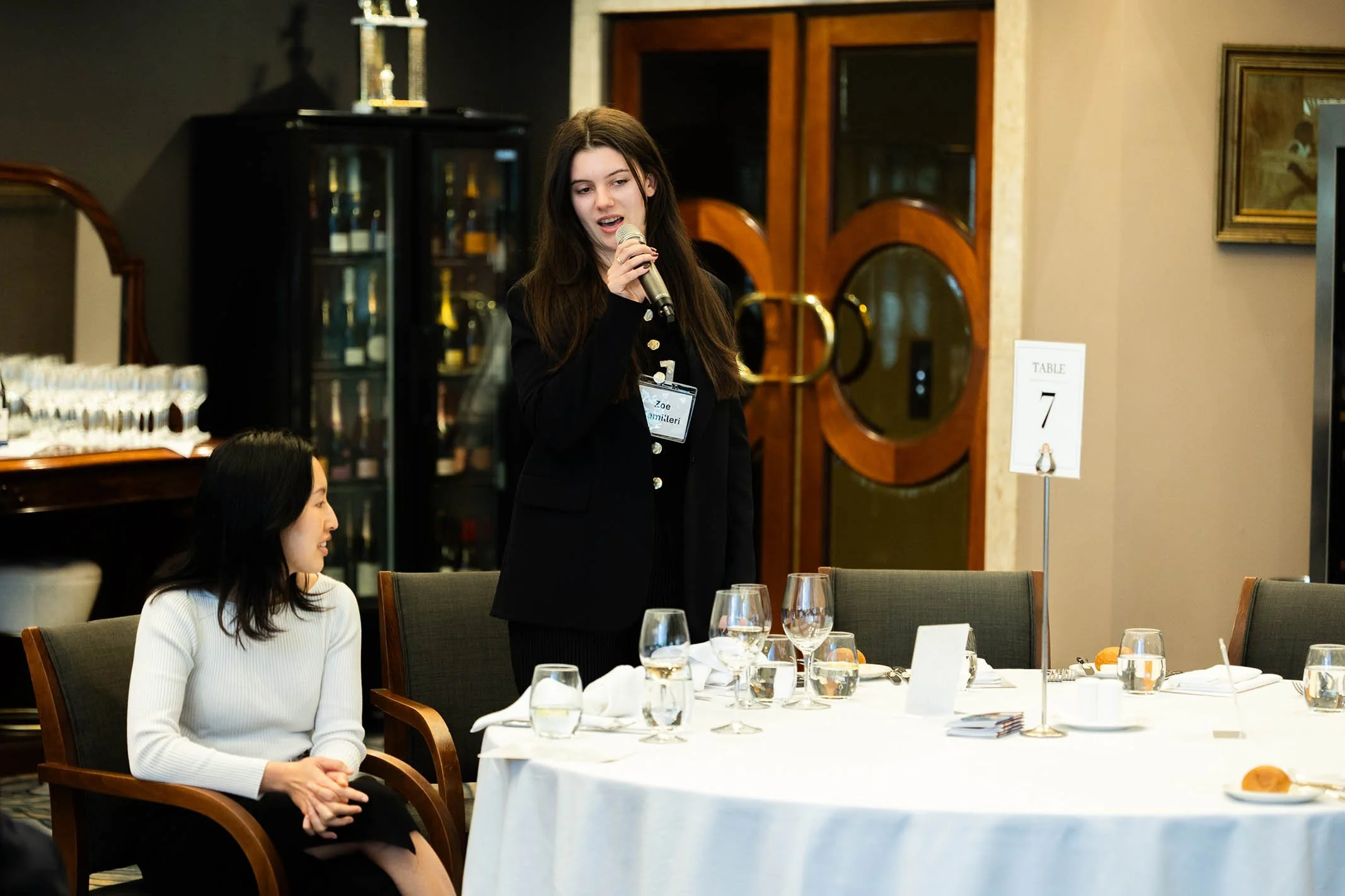 A woman standing and speaking into a microphone at a formal dining event, while another woman sits nearby at a round table set with glasses, plates, and silverware. The setting appears to be a banquet or conference room.