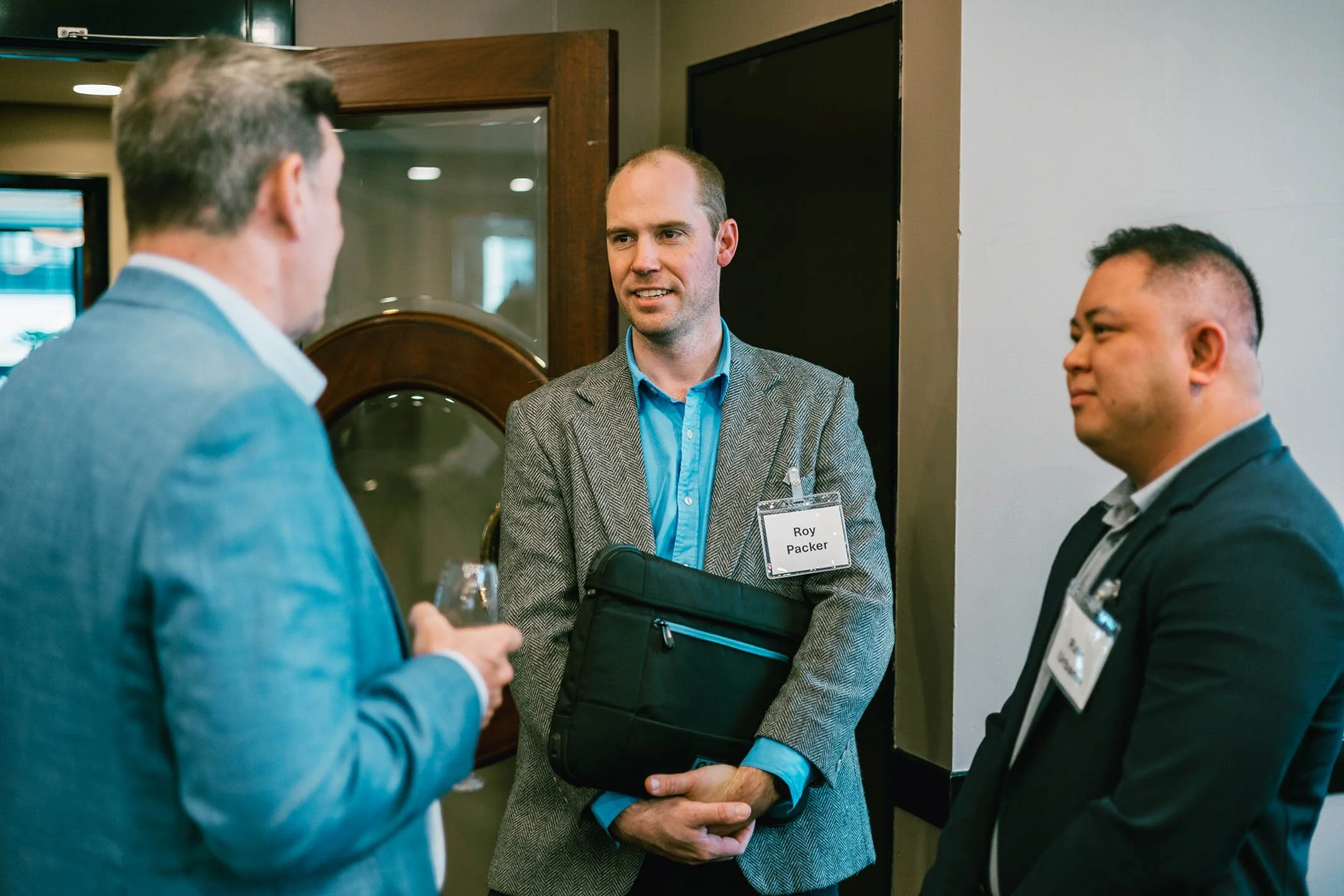 Three men in business attire having a conversation at a professional event. The man in the middle, named Roy Packer, wears a gray blazer and holds a black briefcase. The man on the left is looking at his phone, and the man on the right has short hair