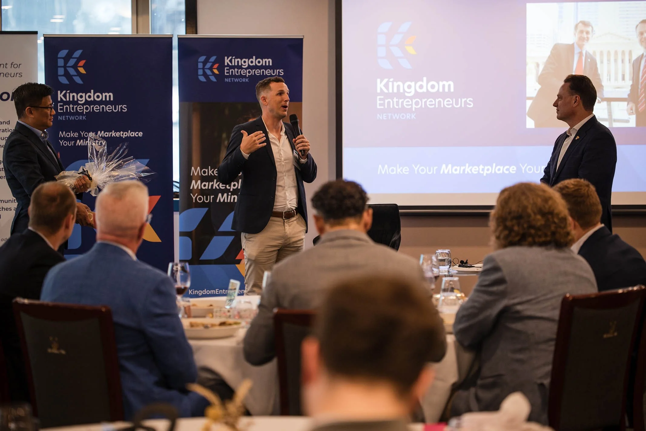 A conference or networking event with several people seated at round tables, a speaker in a dark blazer and light pants speaking into a microphone on stage, and another person holding a bouquet of flowers. A large screen behind shows the 'Kingdom Ent