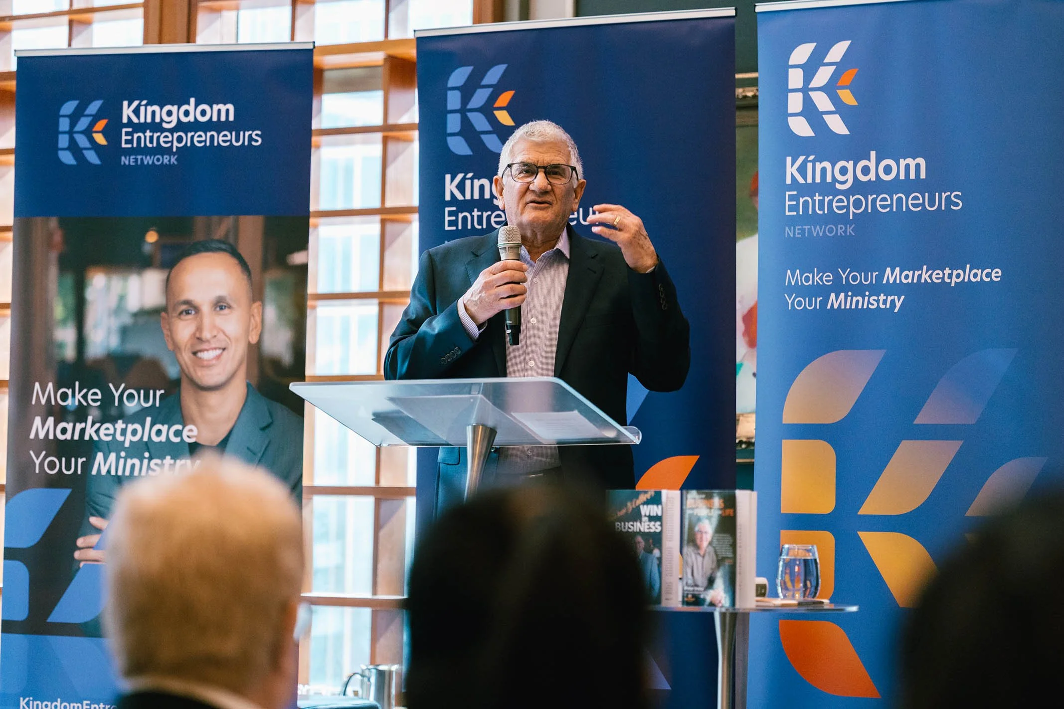 An older man with glasses wearing a suit, speaking into a microphone at a podium during a Kingdom Entrepreneurs Network event. Behind him are two blue banners with the organization's logo and the slogan 'Make Your Marketplace Your Ministry.'