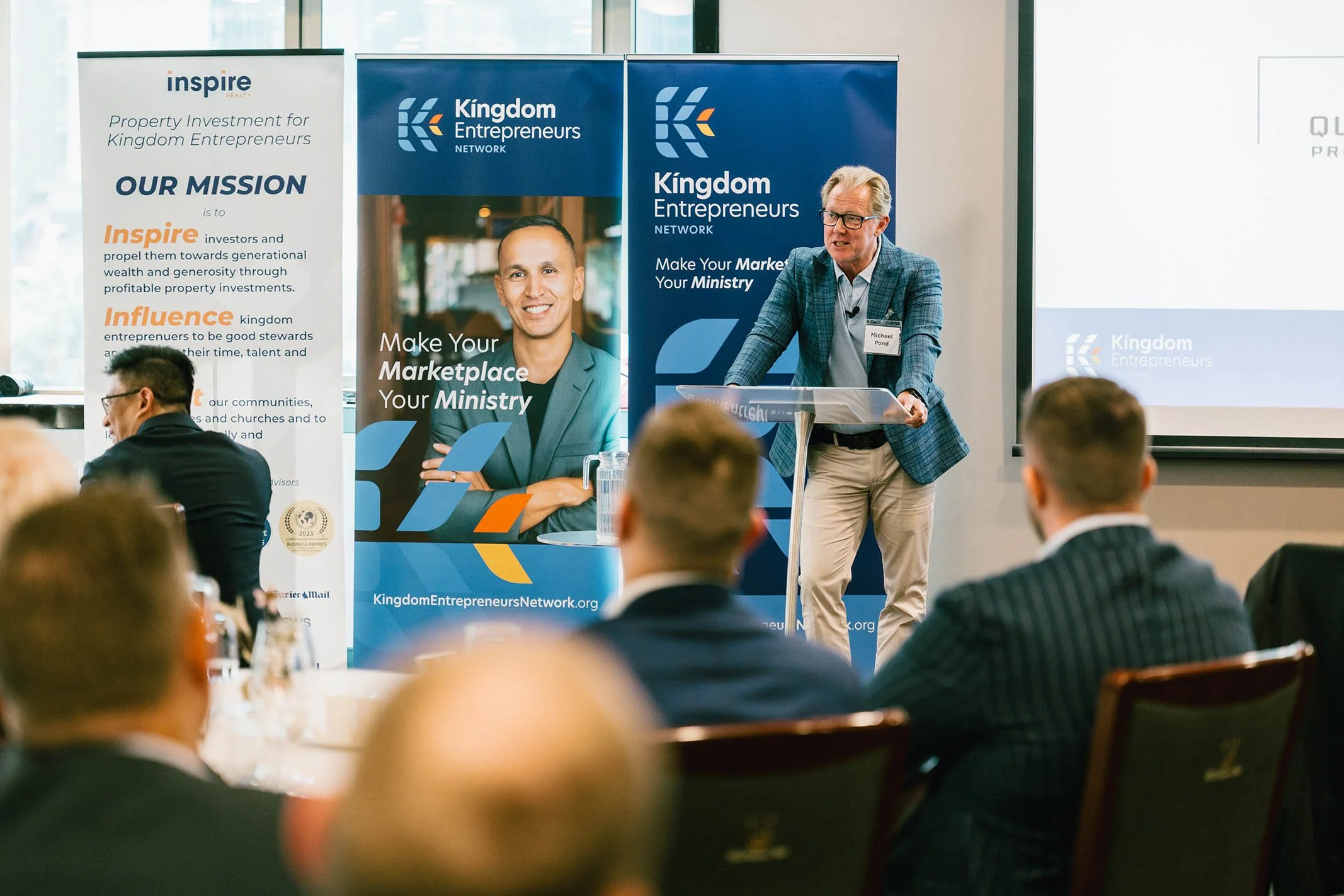 A man in a blue blazer and glasses is standing at a podium speaking to an audience at a conference. Behind him are banners with the logos of Kingdom Entrepreneurs Network and Inspire Realty, along with a group of seated attendees listening. The venue