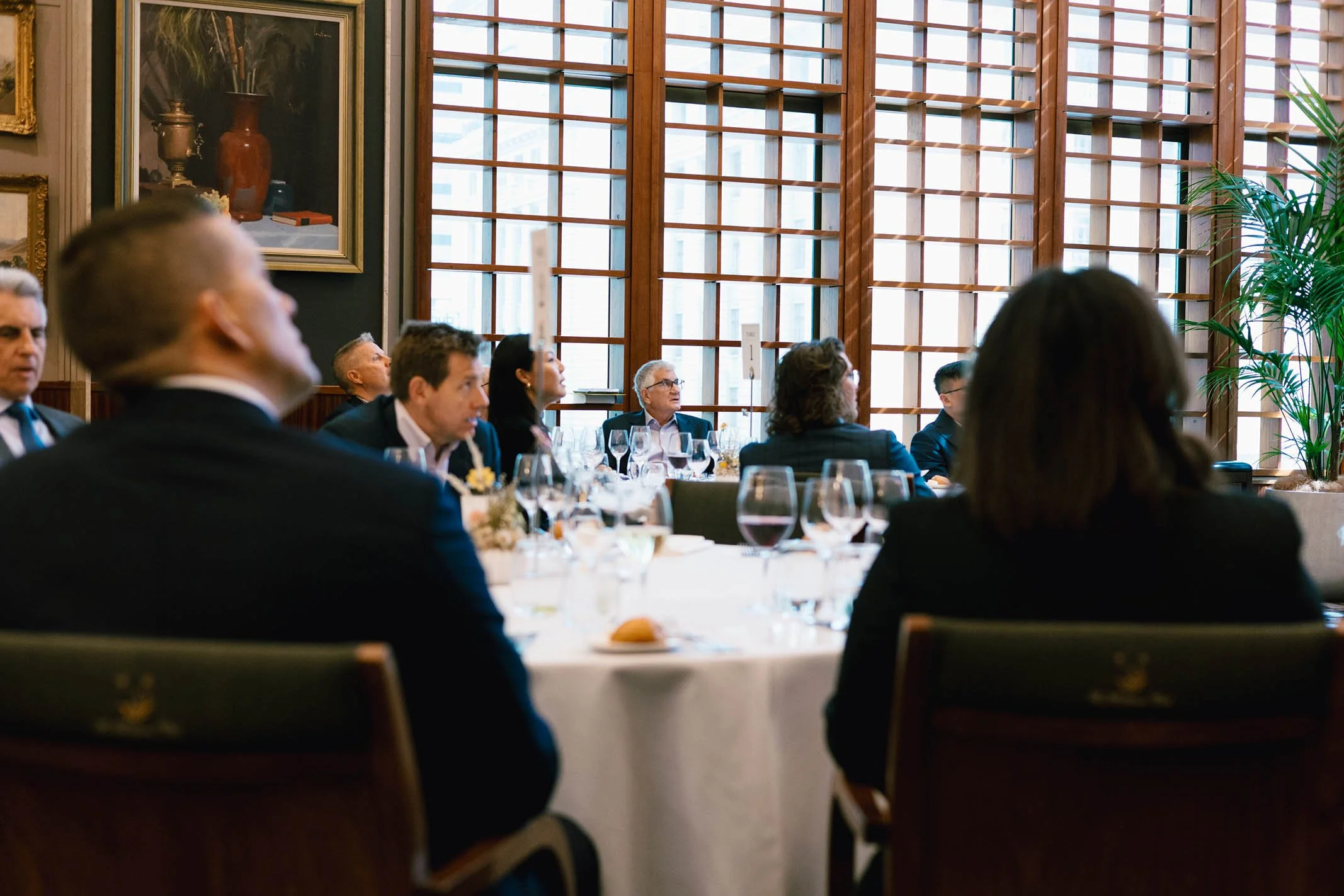 People seated at a formal event, wearing business attire, inside a room with large wooden windows and framed paintings on the wall.