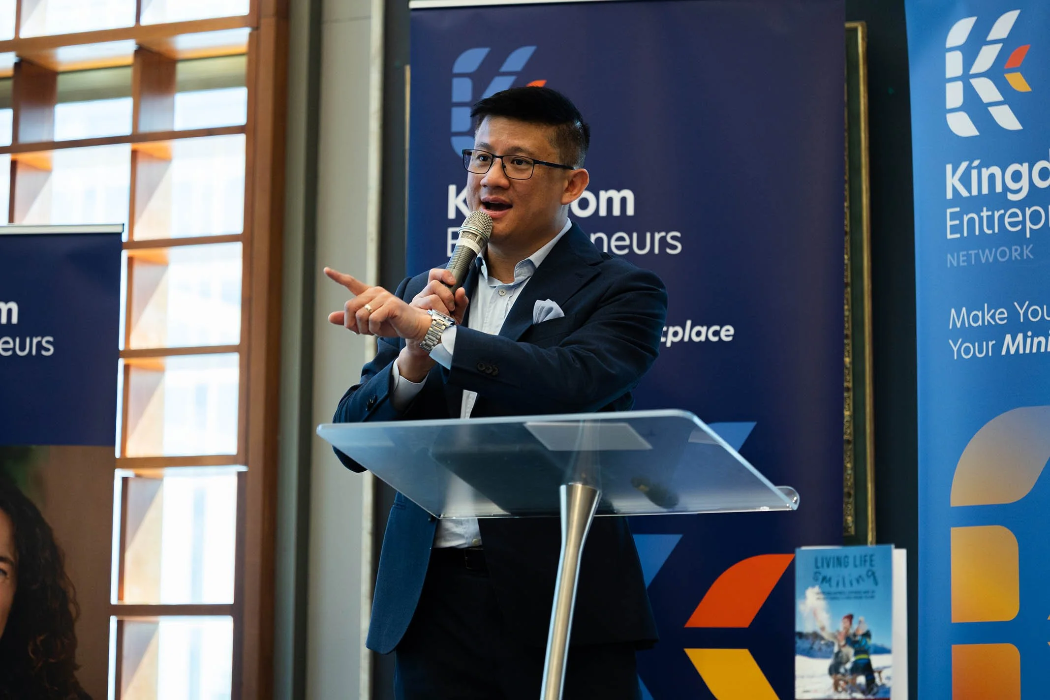 A man in a dark suit speaking into a microphone at a podium during a professional event. Behind him are blue banners with white and orange logos and text related to entrepreneurship.