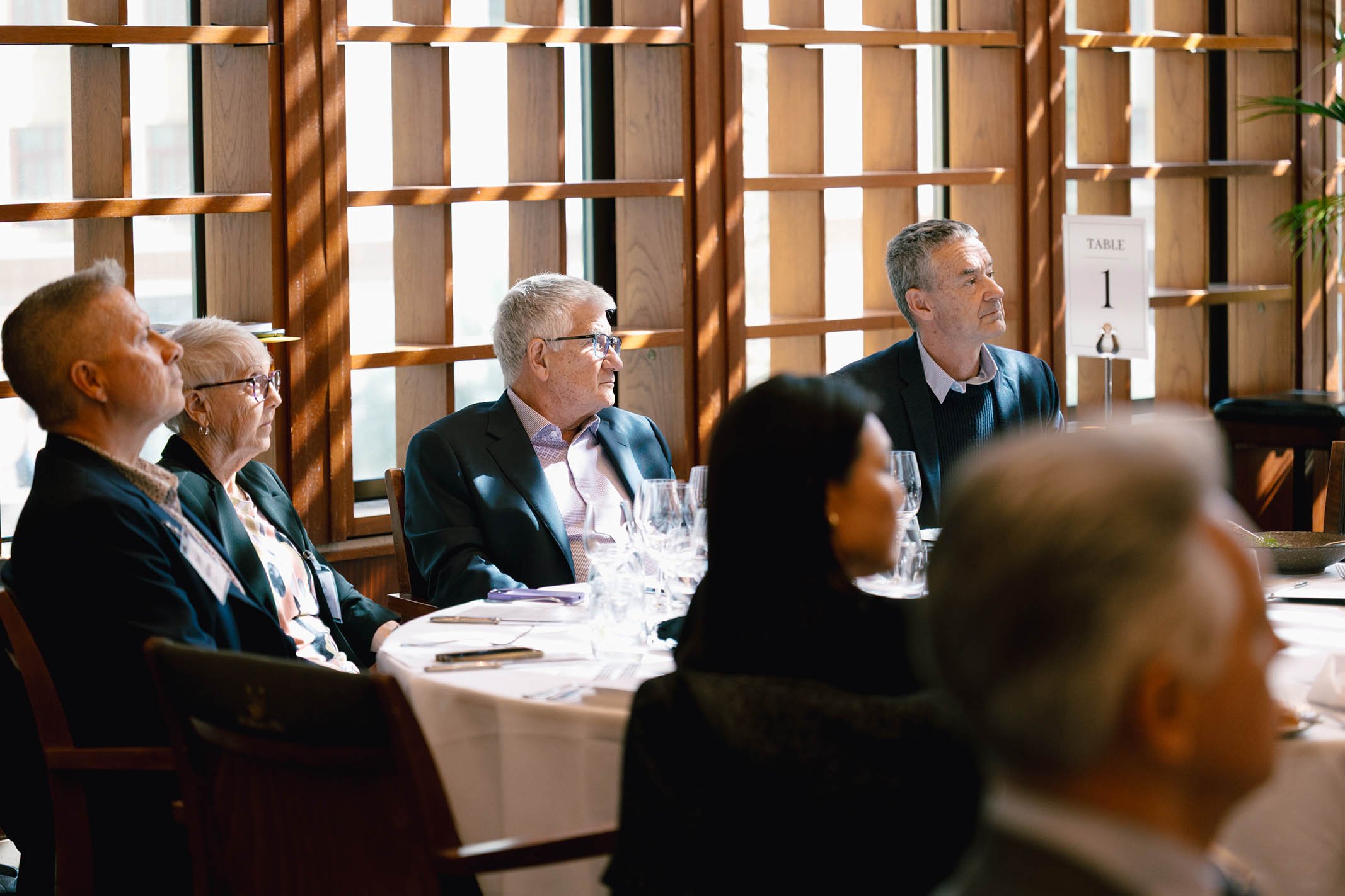 Attendees sitting at a table during a formal event or meeting, listening to a speaker or presentation in a well-lit room with wooden lattice walls and large windows.