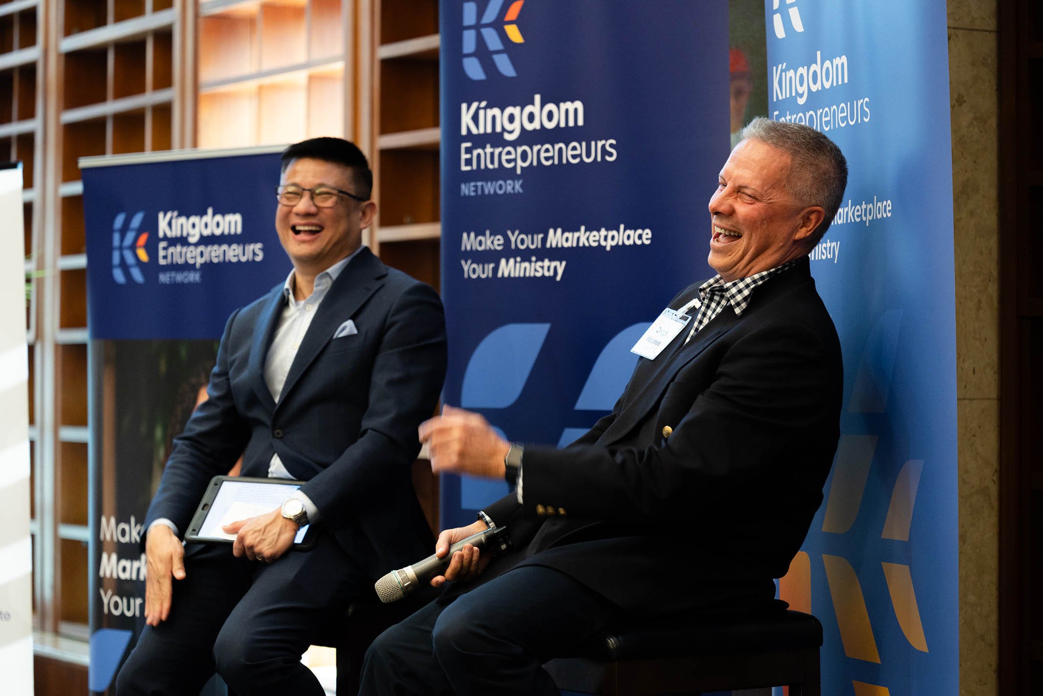 Two men laughing and smiling on stage at Kingdom Entrepreneurs event, sitting in front of blue banners with their logo and text, one holding a tablet and the other a microphone.