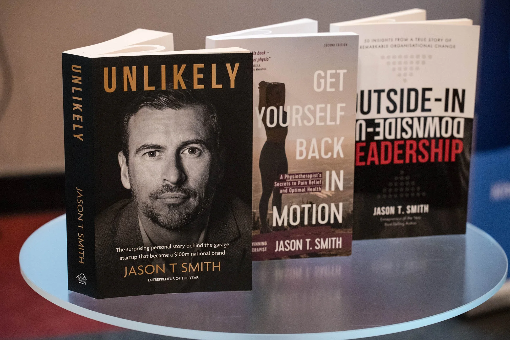 Three books standing on a round table, with the front book titled "Unlikely" by Jason T. Smith, featuring a black-and-white photo of a man's face on the cover.