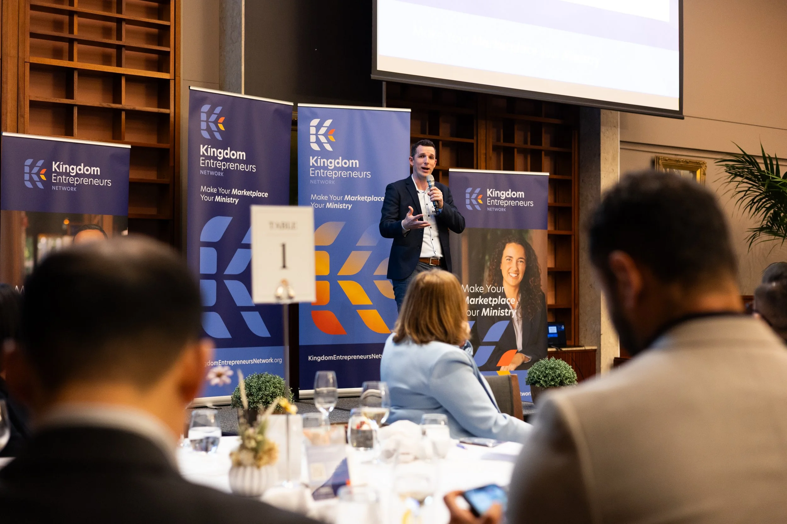 A man in a dark suit speaking at a presentation event with kingdom entrepreneurs banners in the background, seated audience, a woman with long curly hair and a white shirt sitting at a table, table number 1 sign, and a large screen above the speaker.