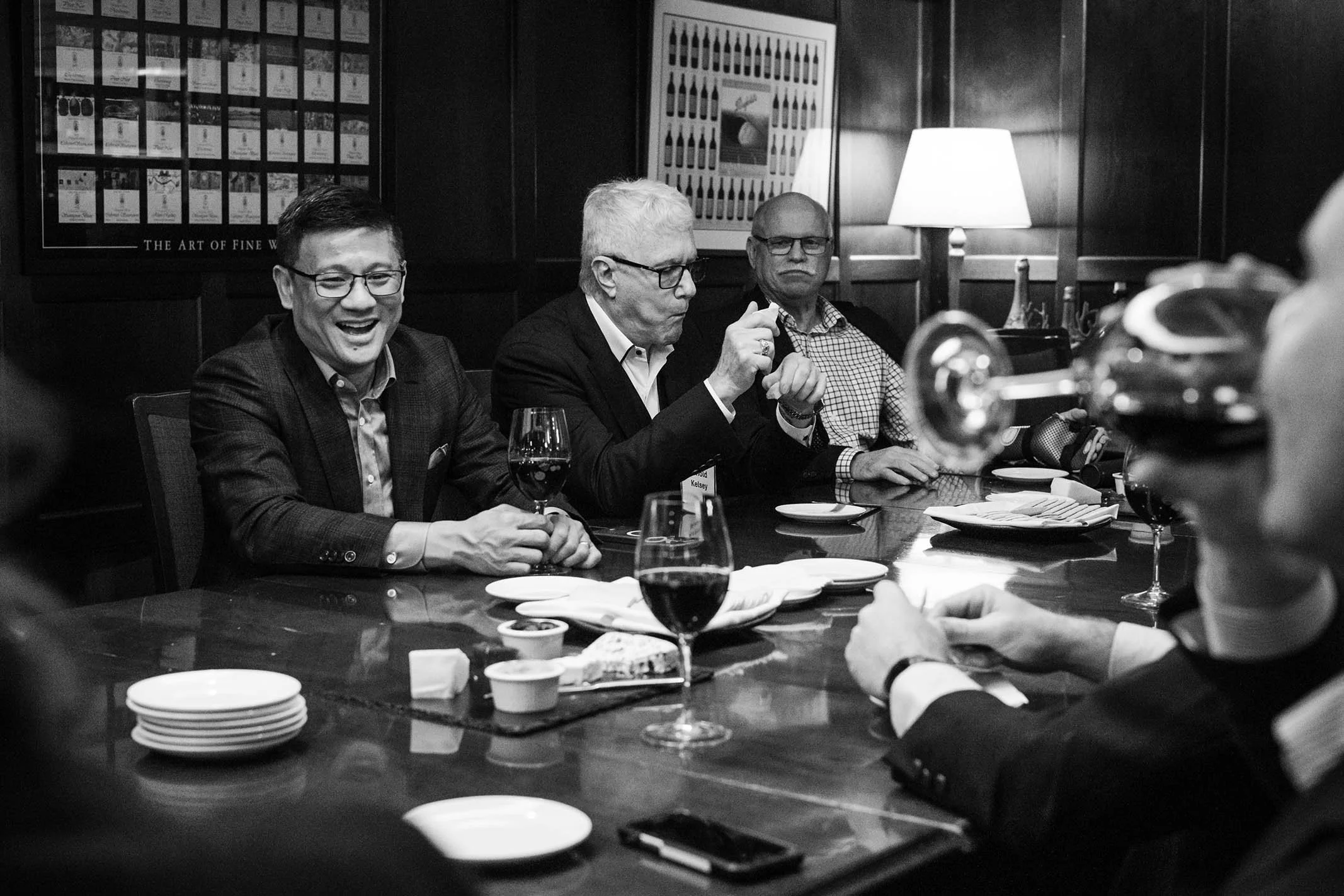A group of men sitting around a dinner table engaged in conversation, with wine glasses, plates, and small dishes on the table, in a dimly lit restaurant or private room.