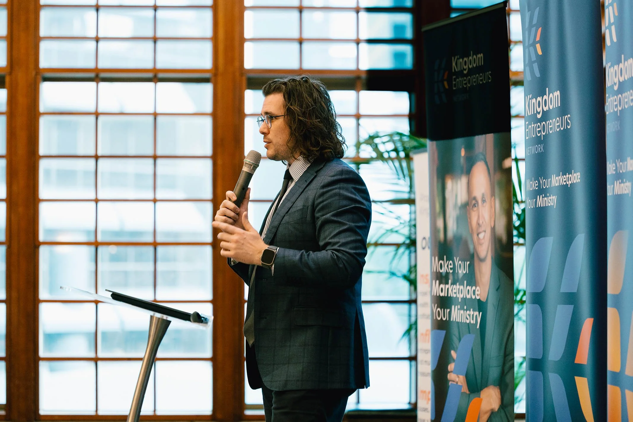 A man with glasses and shoulder-length hair in a suit, holding a microphone and speaking at a conference with banners behind him that read 'Kingdom Entrepreneurs' and 'Make Your Marketplace Your Ministry'. The background features large windows with w