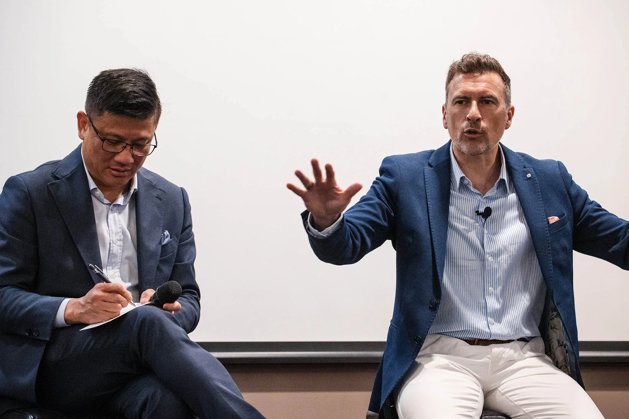 Two men in business attire are engaged in a discussion; the man on the right is speaking with his hand raised, while the man on the left is writing notes.