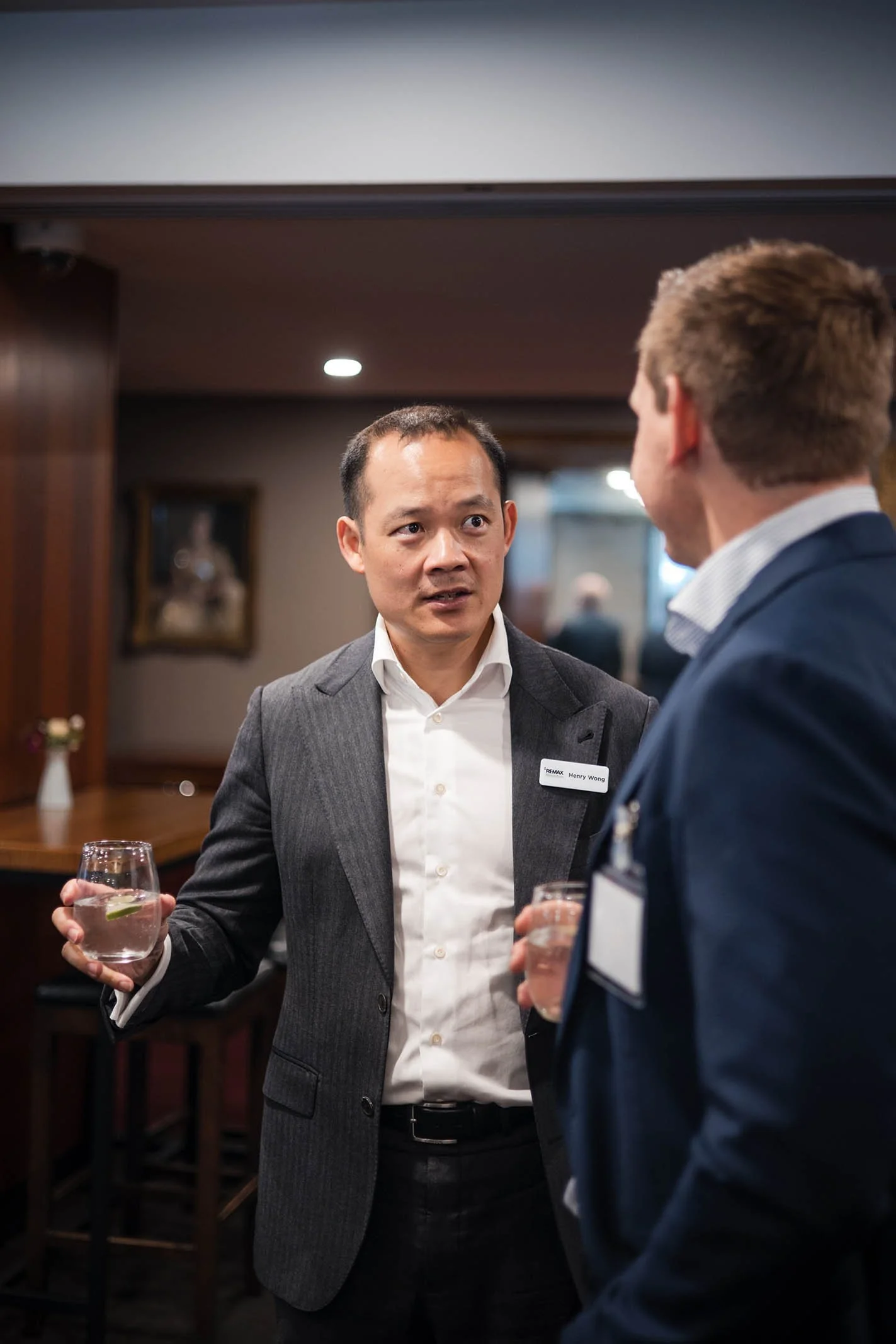 Two men in business attire engaged in conversation at a formal event, holding glasses of water, in an indoor setting with warm lighting and framed artwork on the wall.