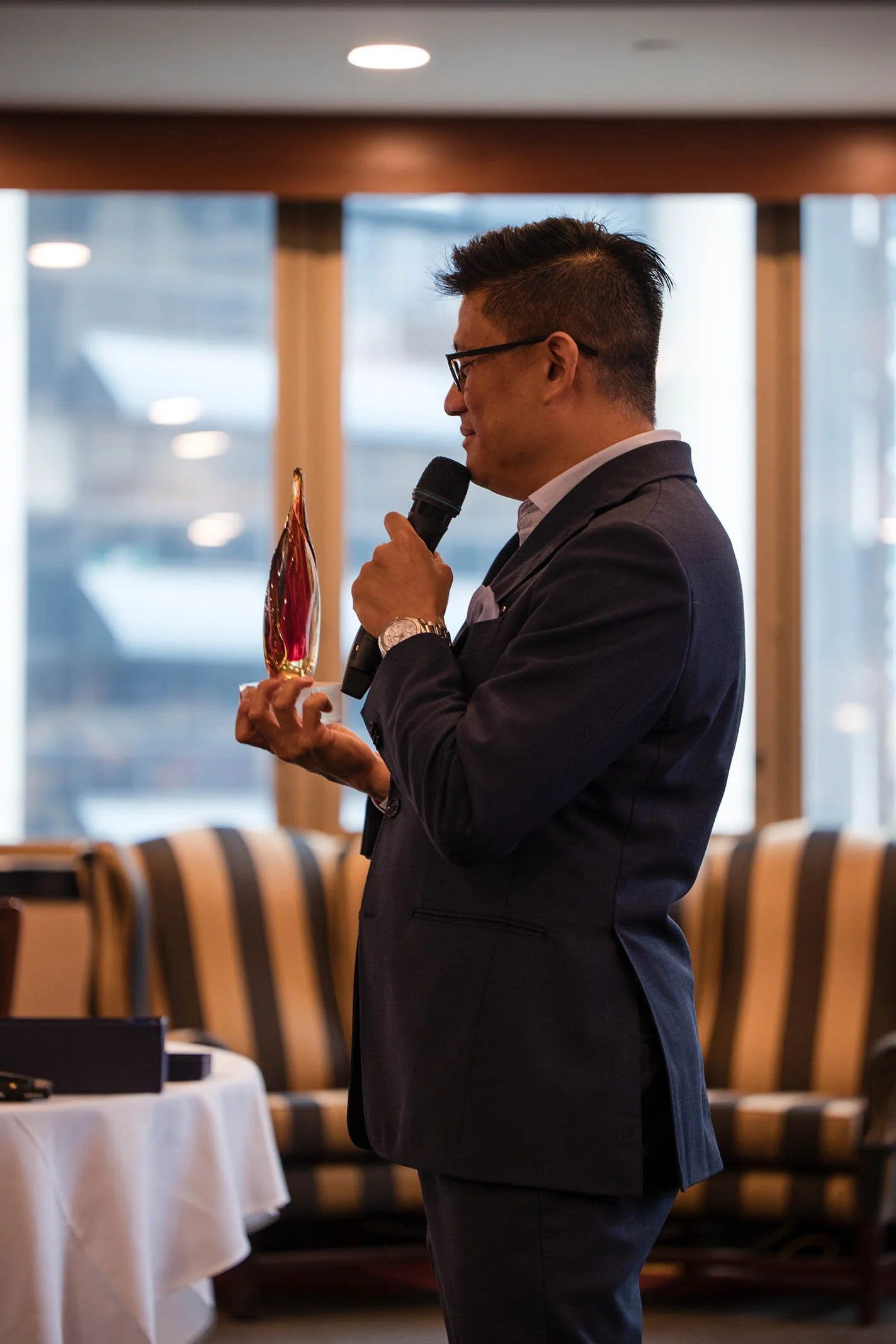 Man in suit holding an award and speaking into a microphone at an indoor event.