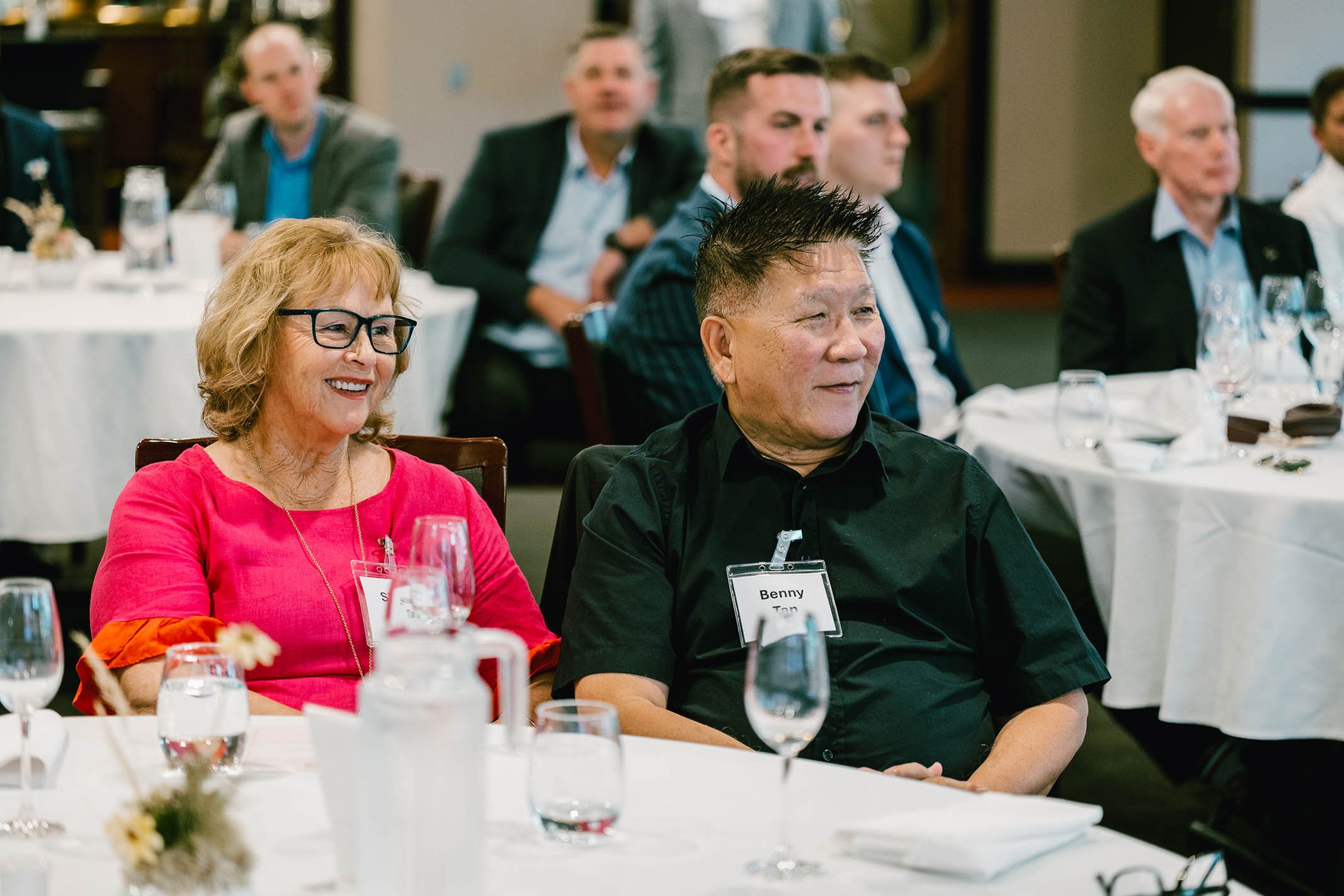 A group of people sitting at a banquet table, listening attentively at a formal event. In the foreground, a woman wearing glasses and a pink top smiles, next to a man with a name tag that reads 'Benny Tan' in a black shirt. Several other attendees ar