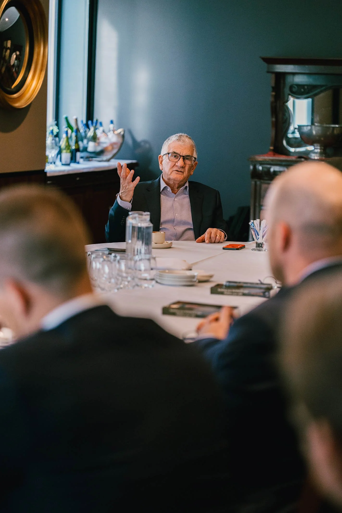 Old man in glasses and a suit speaking or giving a presentation during a business meeting in a conference room with several attendees.