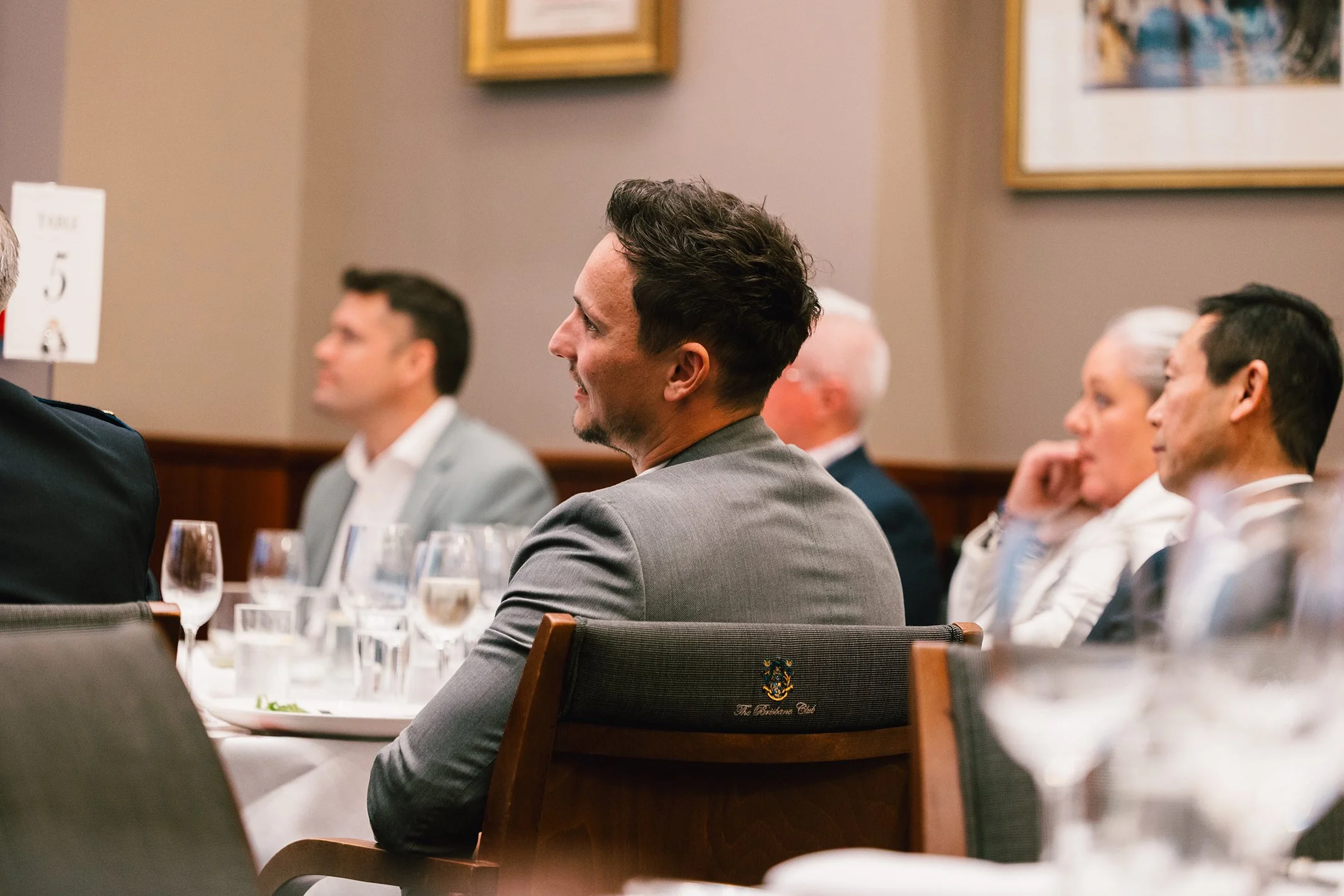 People in business attire attending a formal event or conference, sitting at tables with wine glasses and water glasses, in a room with framed artwork on the wall.