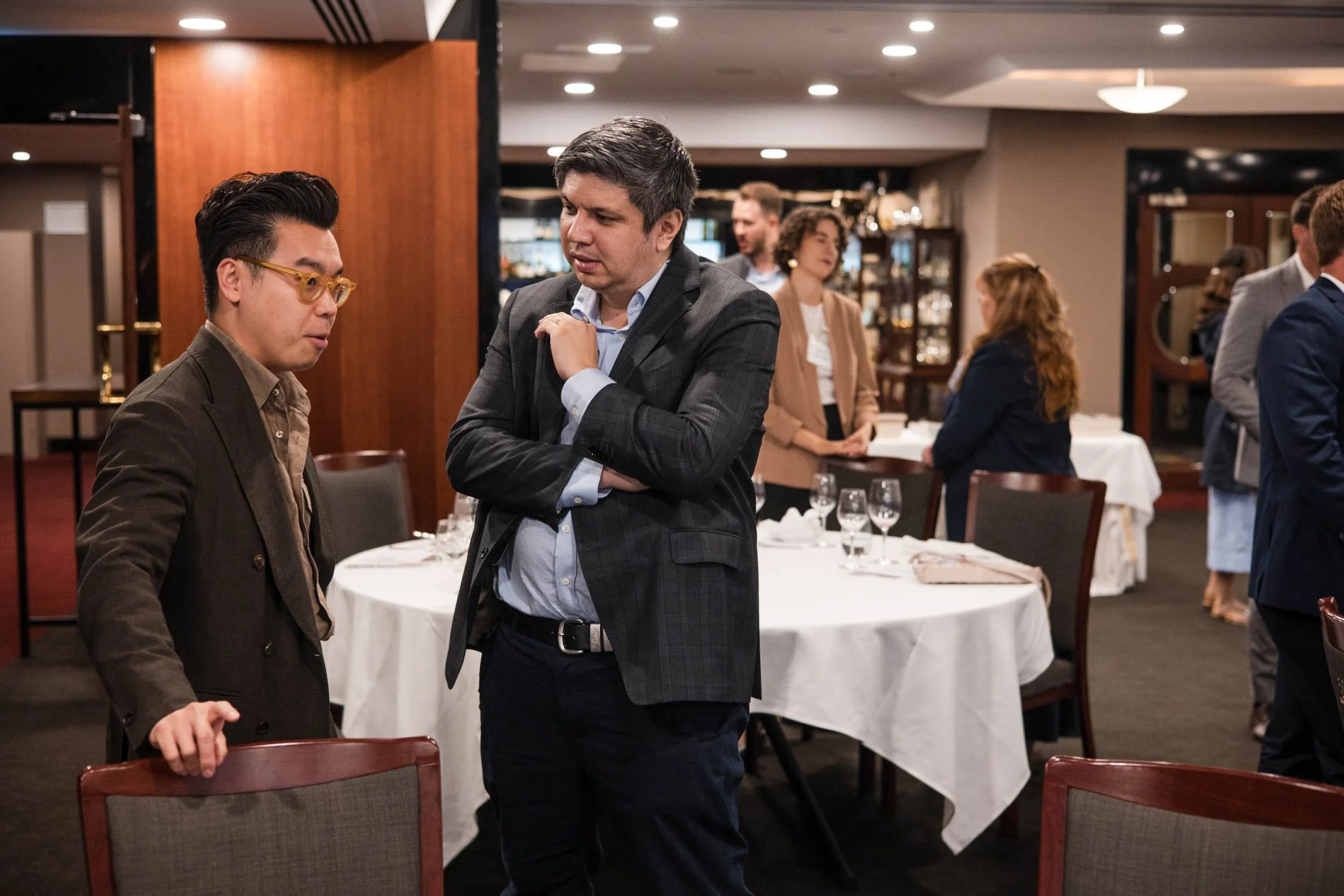 Two men engaged in conversation at a formal event in a restaurant or banquet hall, with tables set with white tablecloths, glasses, and silverware, and other attendees in the background.