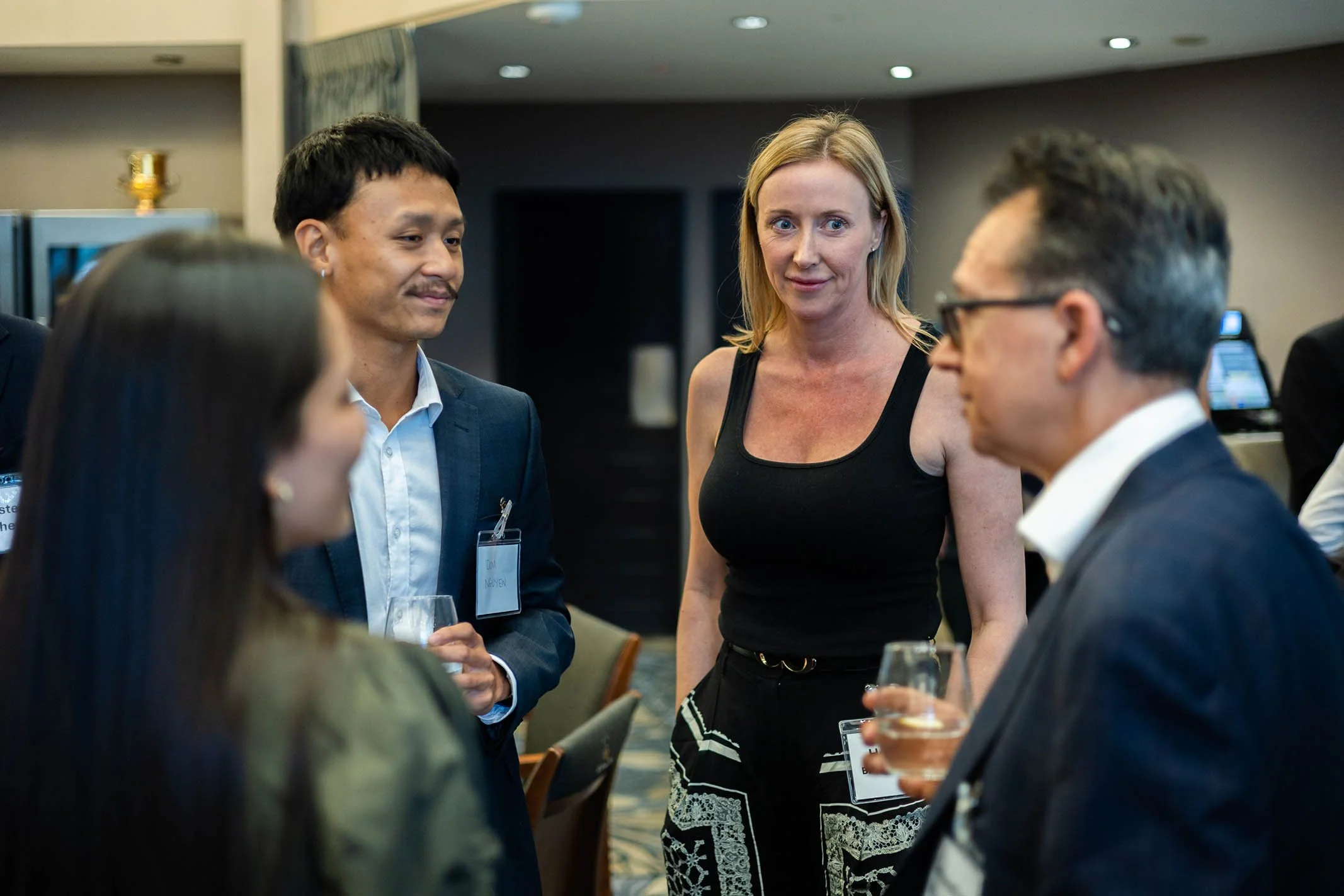 A group of people engaged in conversation at a professional event, with one woman in a black tank top and patterned pants talking to a man in glasses and a suit, while two others are partially visible.