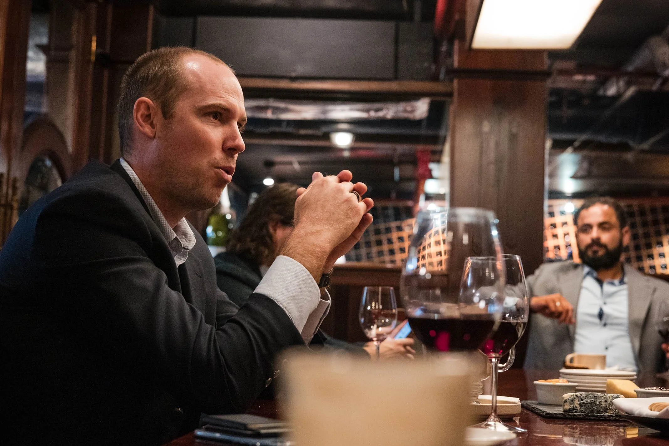 A group of people sitting around a dinner table in a dimly lit restaurant, with two glasses of red wine visible in the foreground, and a man in a gray blazer listening attentively.