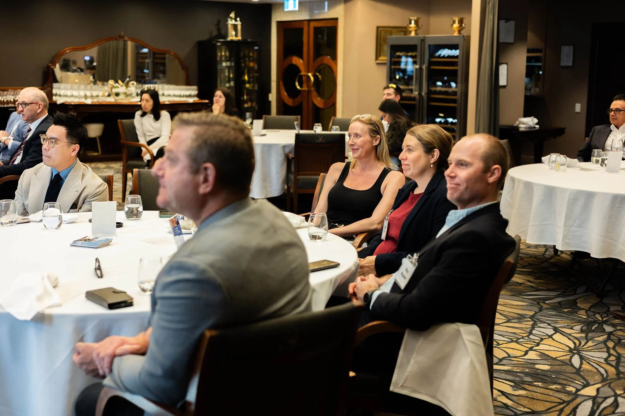 People attending a formal event or conference sitting at round tables with white tablecloths, listening attentively, some smiling in a well-decorated room with wooden furniture, glass display cabinets, and artwork on the walls.