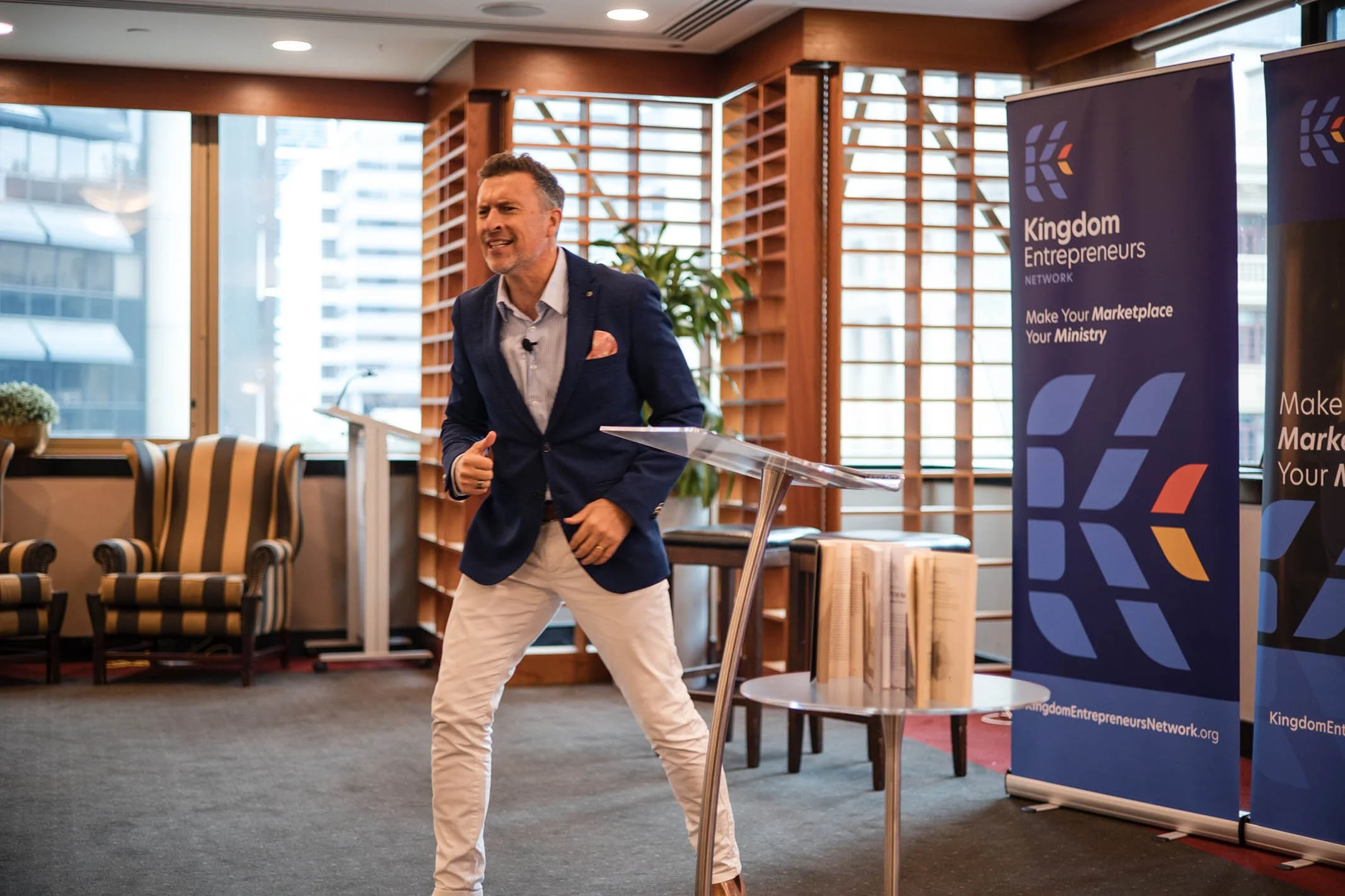 Man giving a presentation at a conference, standing at a transparent podium, with banners displaying 'Kingdom Entrepreneurs Network' in the background.