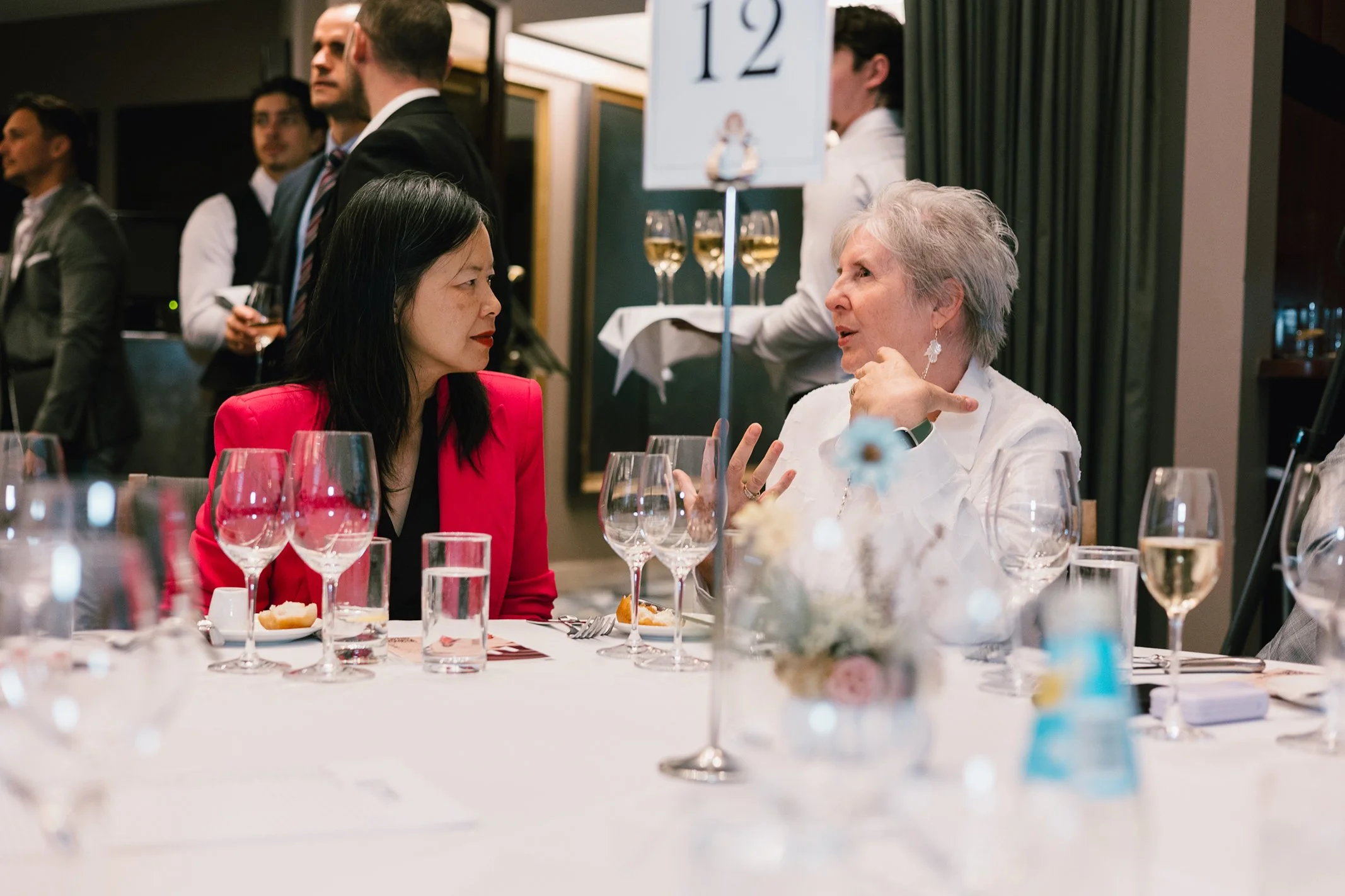 Two women engaged in conversation at a formal dinner, surrounded by glasses of wine and wine bottles, with waitstaff in the background and a sign with the number 12 on it.