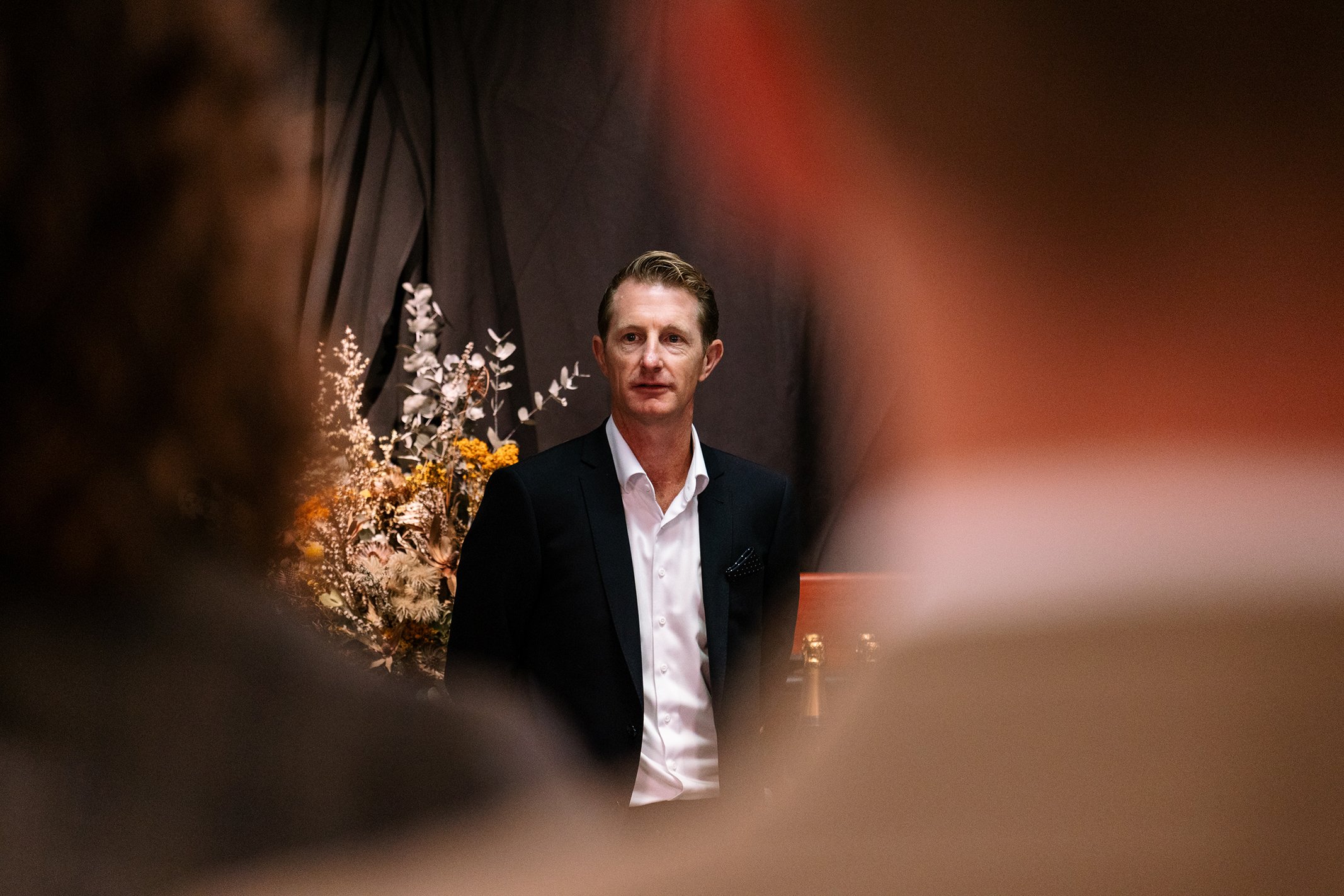 A man in a black suit and white shirt speaking or listening at an event with floral decorations in the background.