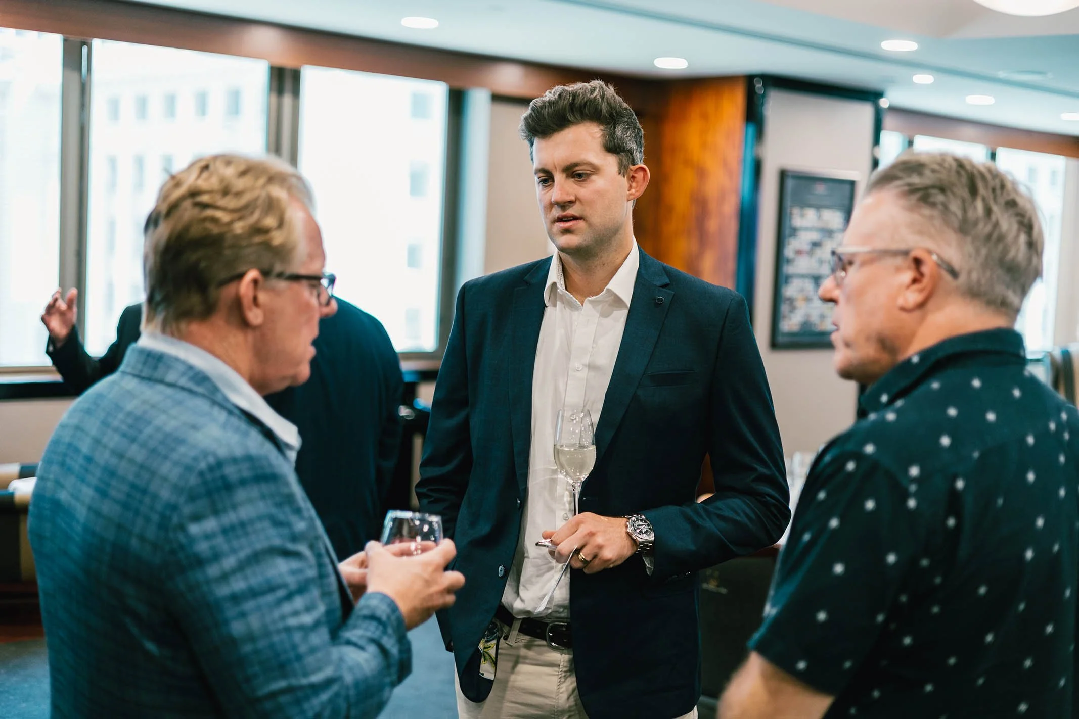 Three men engaged in conversation at a social event, with drinks in hand, indoors with windows and cityscape visible in the background.