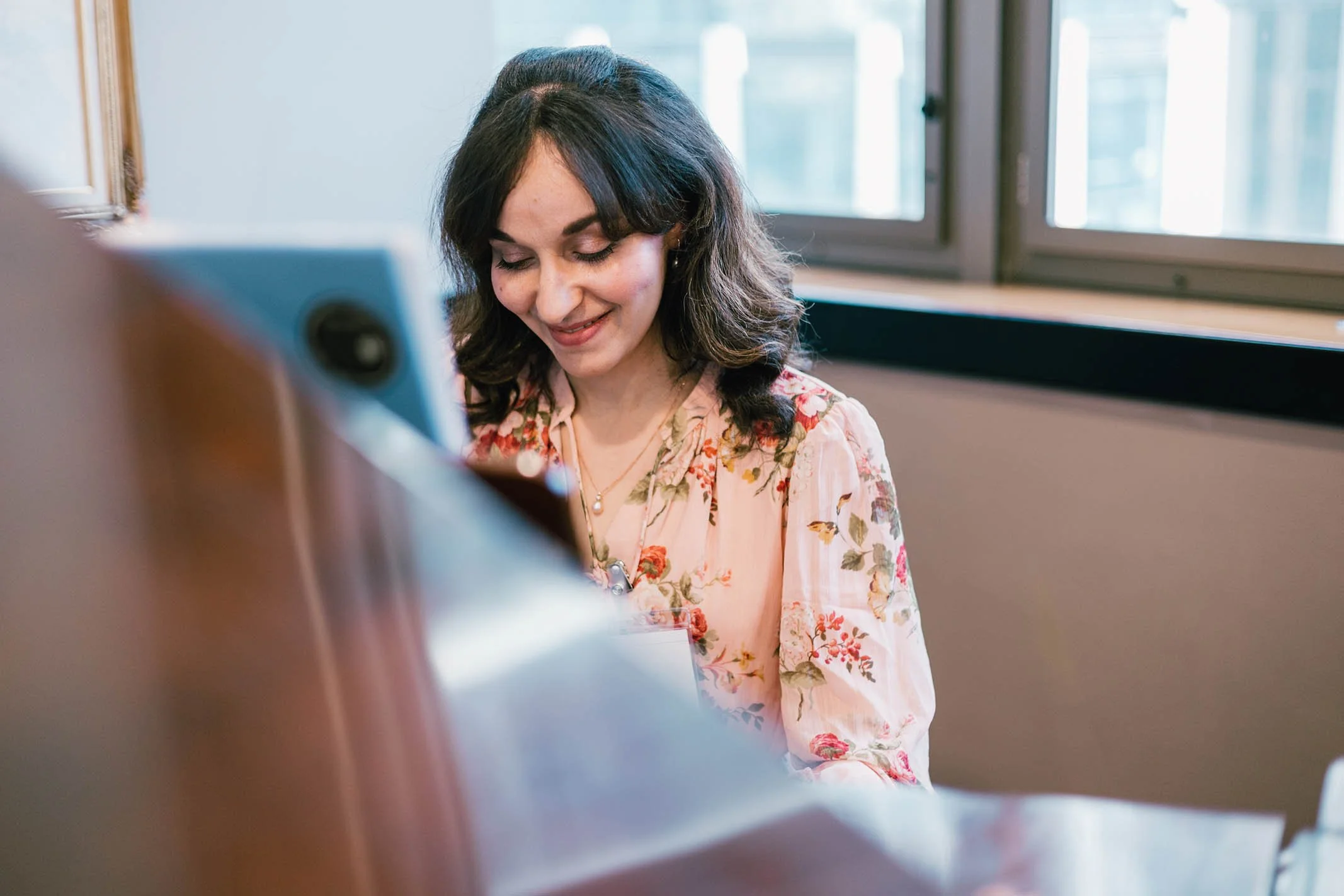 A young woman with dark, wavy hair and light skin, smiling while playing a piano in a room with windows.