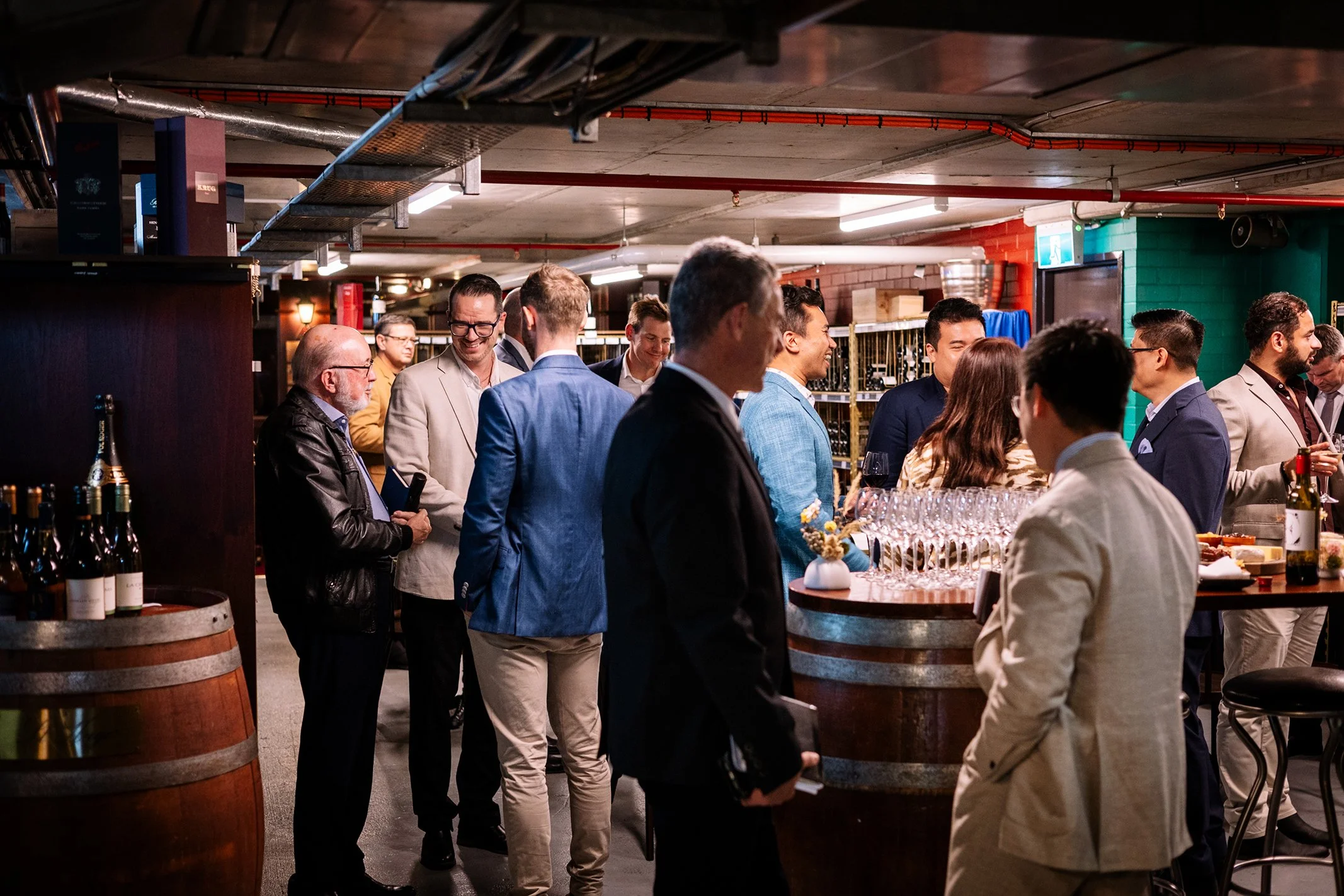 Group of people socializing at a bar in a modern underground venue, some holding glasses of wine, with wine bottles on a barrel in the foreground.