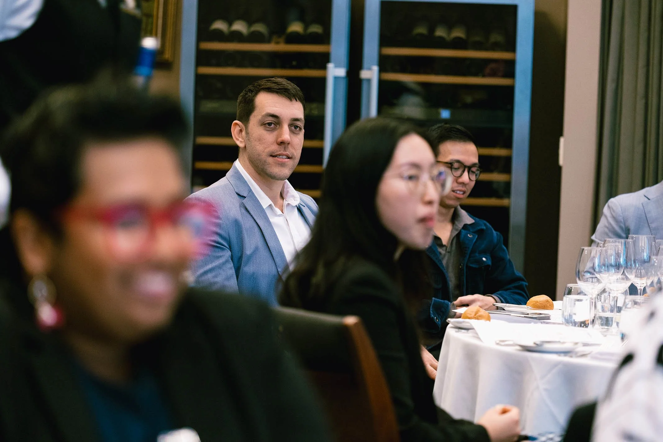 People sitting at a formal dinner table with glasses and bread rolls, possibly at a business or social event.