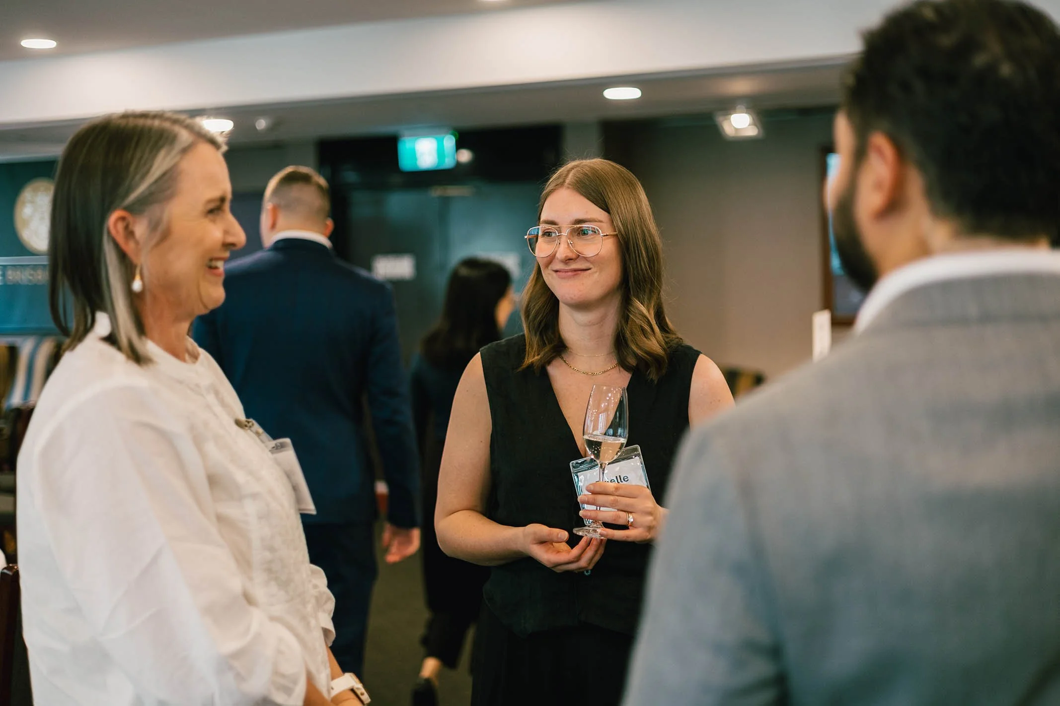 Three people are conversing at a formal event, with one woman holding a glass of rosé wine, and all smiling.