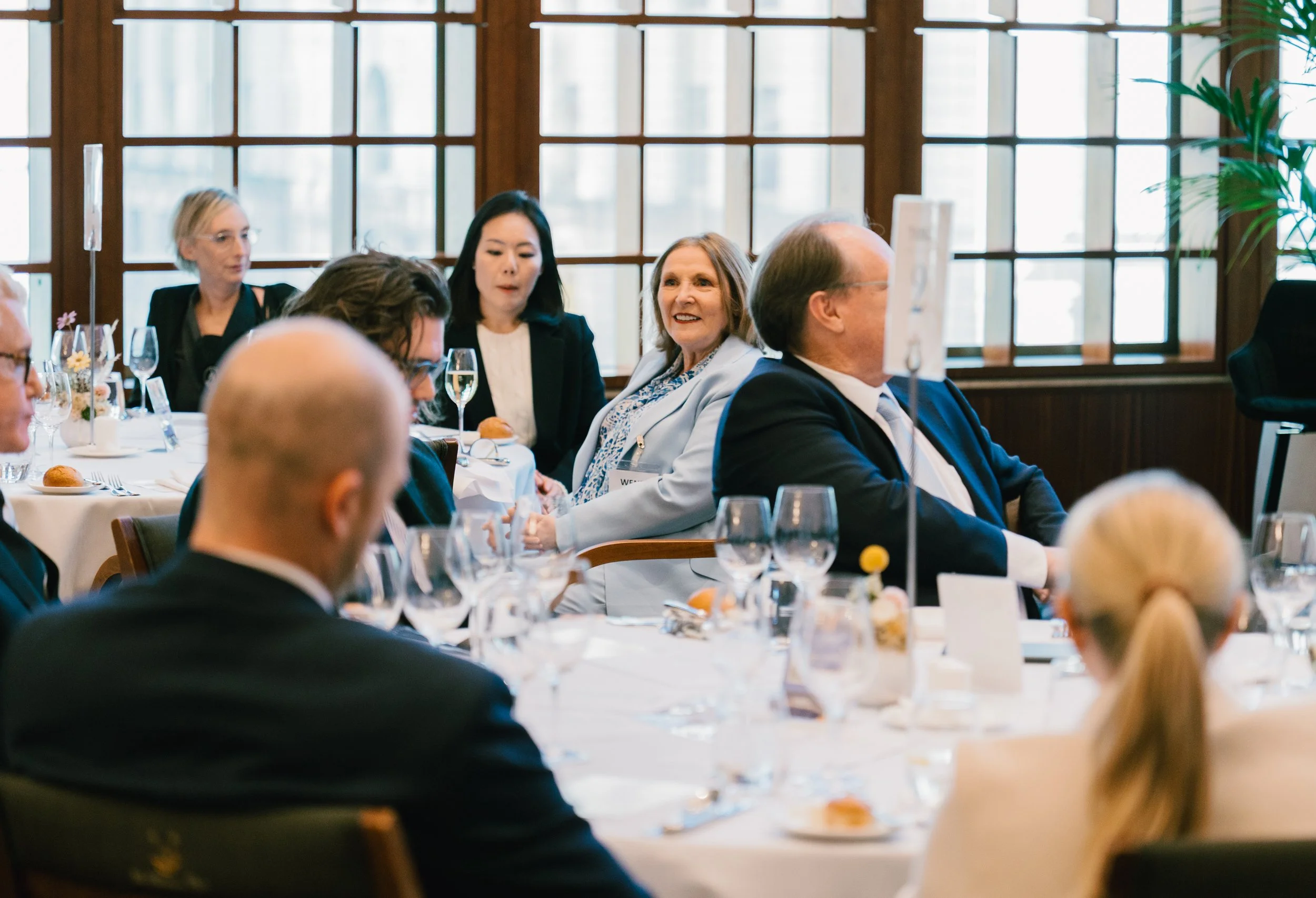 A group of people sitting at a round table during a formal meeting or event, with glasses and tableware visible, in a room with large windows.