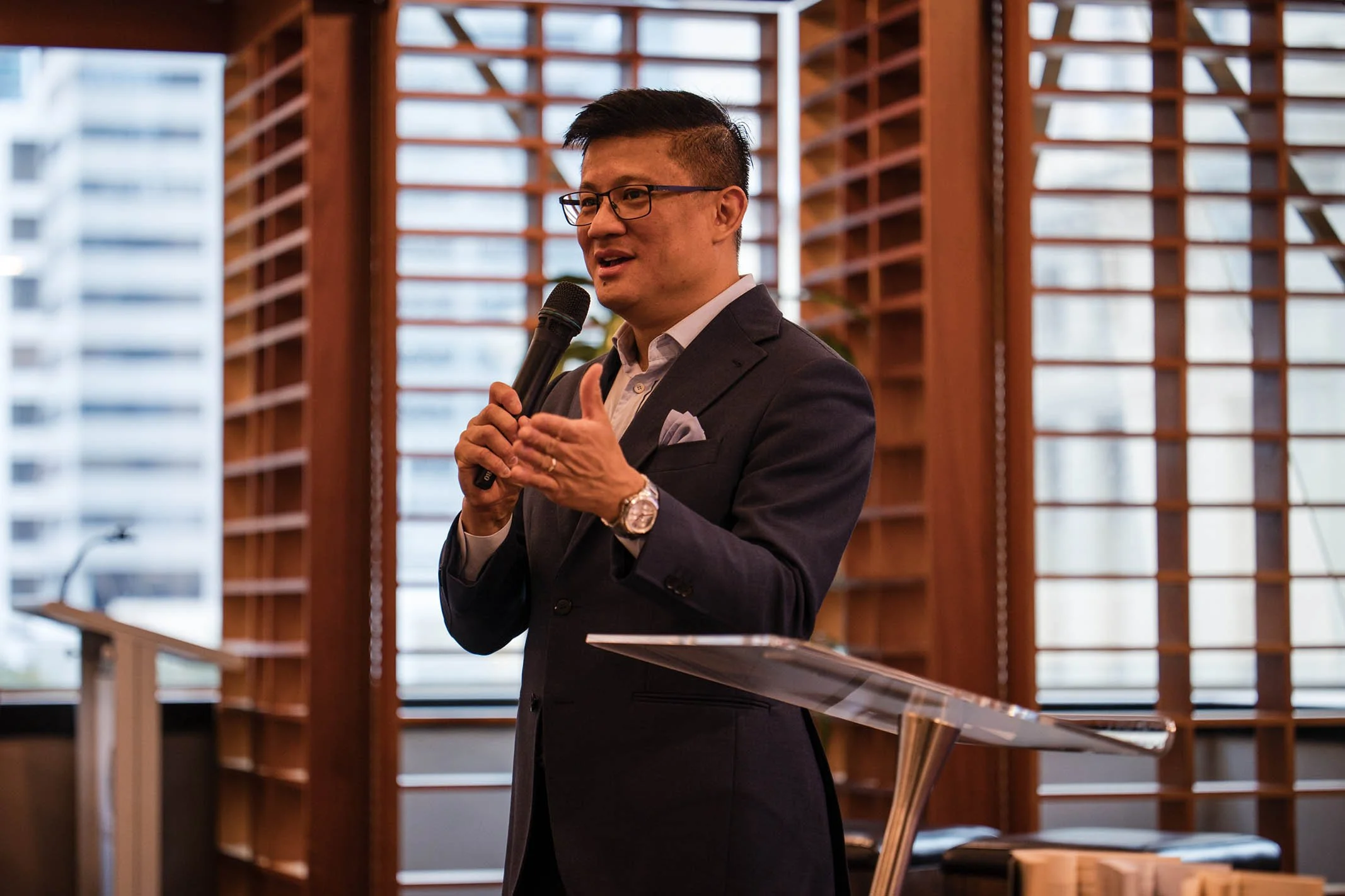 A man in a dark suit and glasses is holding a microphone and speaking at a table in a room with wooden walls and large windows.