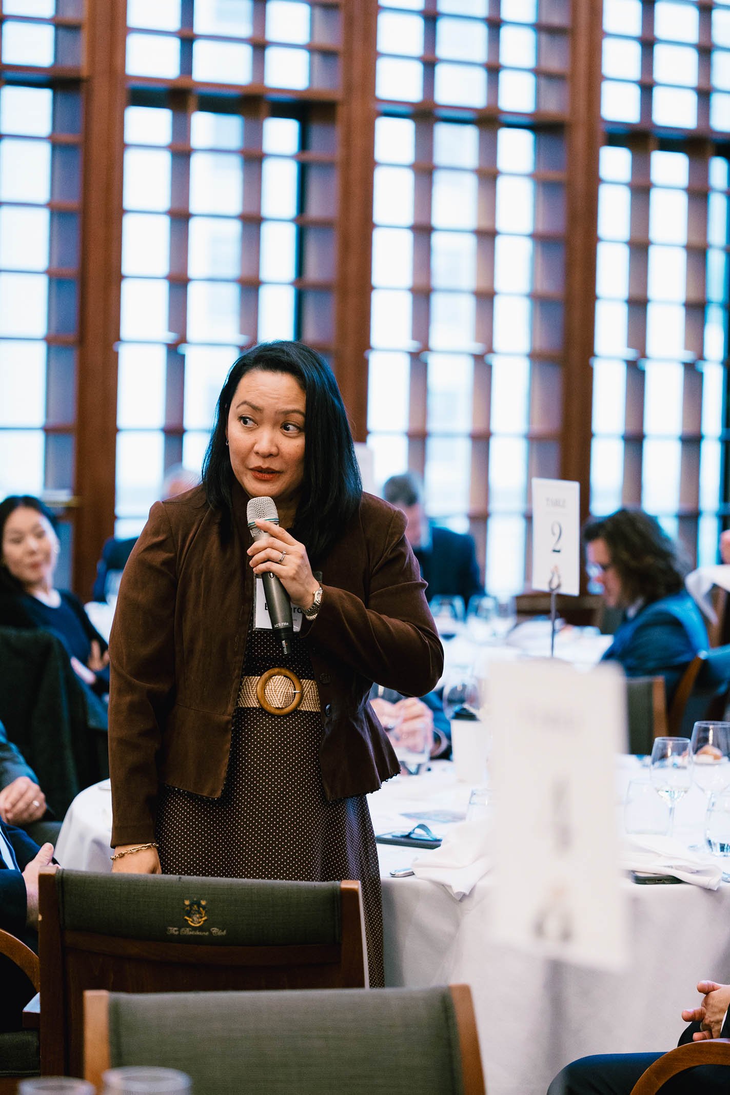 A woman holding a microphone speaks at a formal event in a large room with floor-to-ceiling windows and wooden framing. Several seated attendees are visible, and tables are set with white tablecloths, glassware, and table numbers.