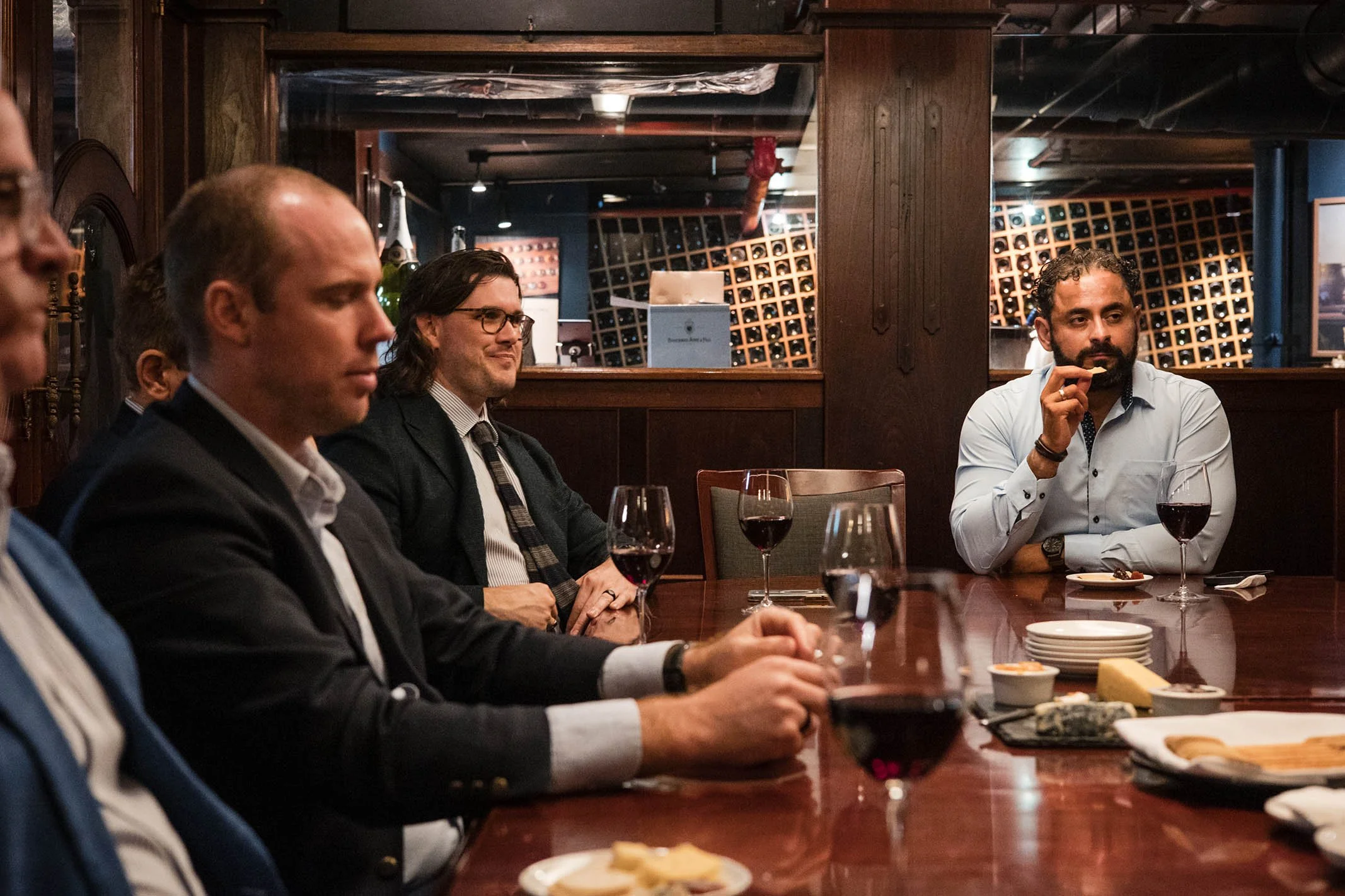 A group of men in suits sitting at a long wooden table in a restaurant or wine cellar, with glasses of red wine and food on the table, engaged in a conversation or discussion.