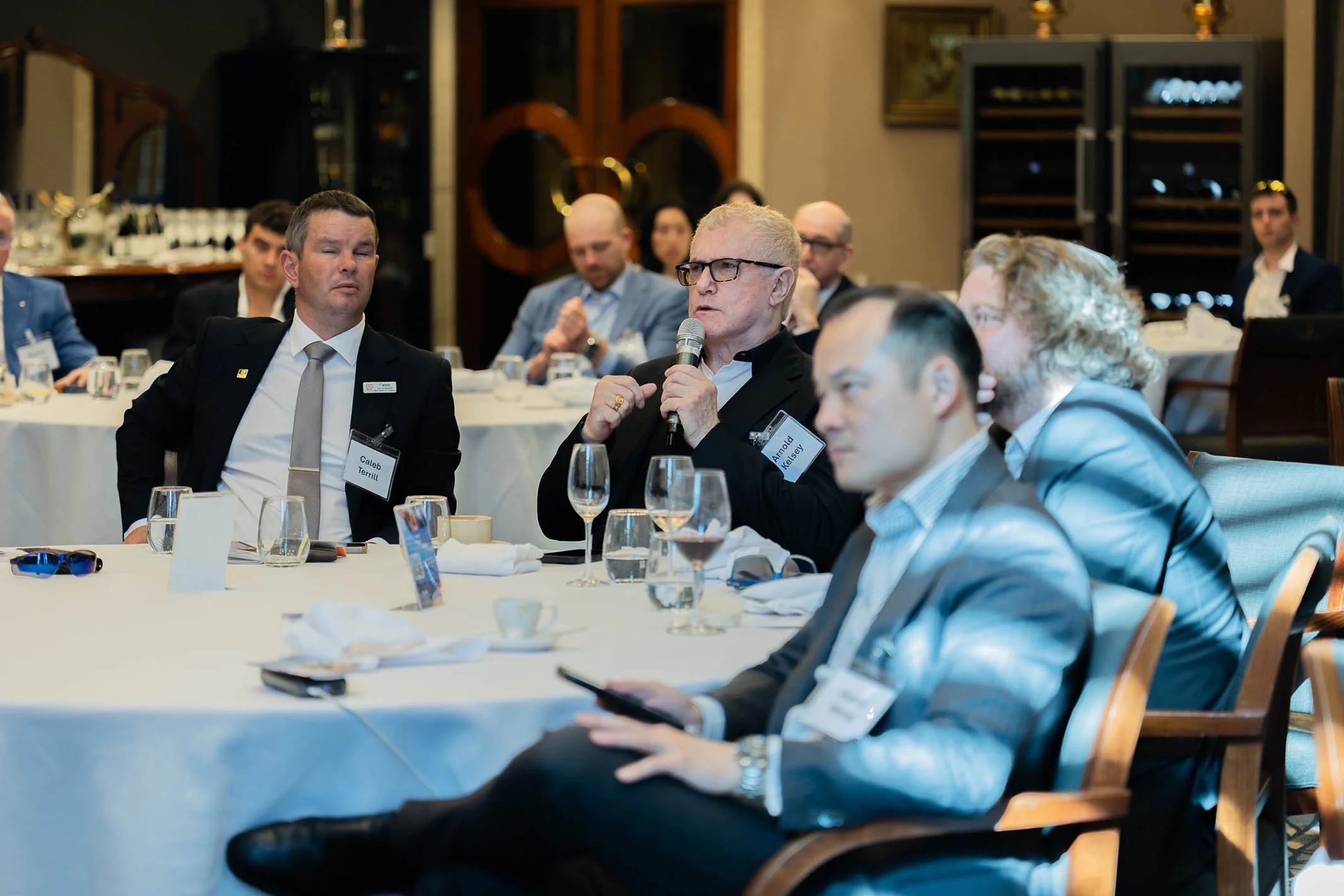A man in a black suit and glasses is speaking into a microphone at a business event, seated at a round table with other professionally dressed attendees, some with name tags, in a formal dining room.