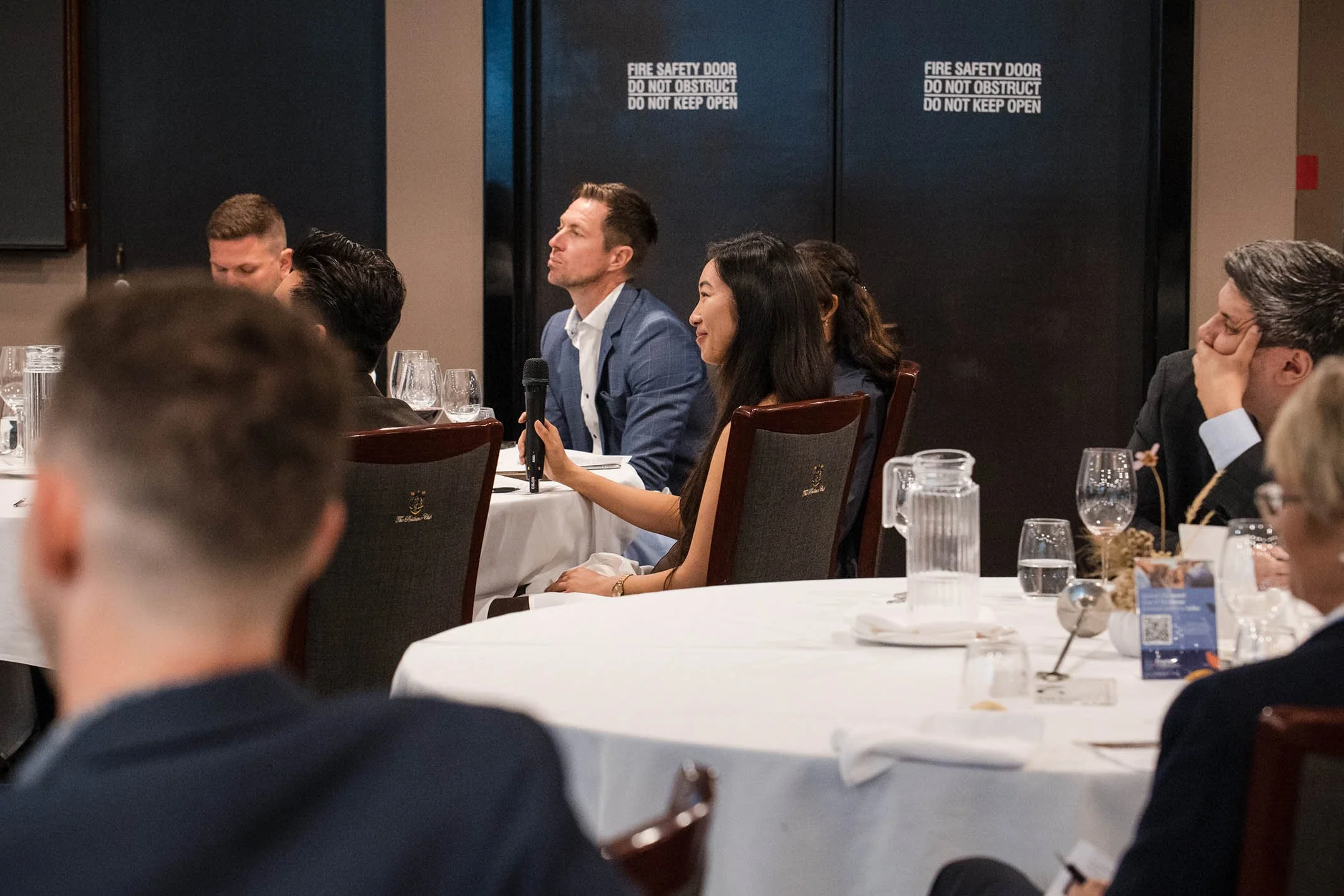 A woman holding a microphone is speaking at a formal event or conference, sitting at a round table with several other attendees, some of whom are listening attentively, with a dark wooden door and glassware on the table in the background.