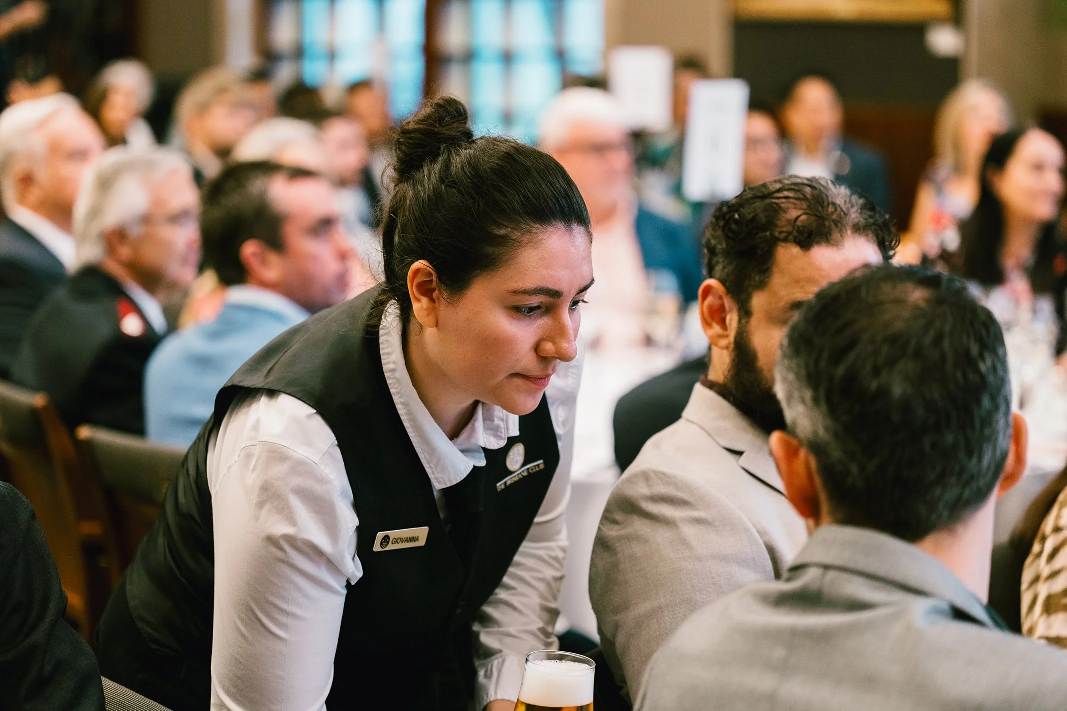 A woman with dark hair tied up, wearing a white shirt and a black vest, leaning forward at a formal event, with a name tag that reads 'Giovanna', holding a glass of beer. Other people are seated around her, paying attention, in a well-lit room with l