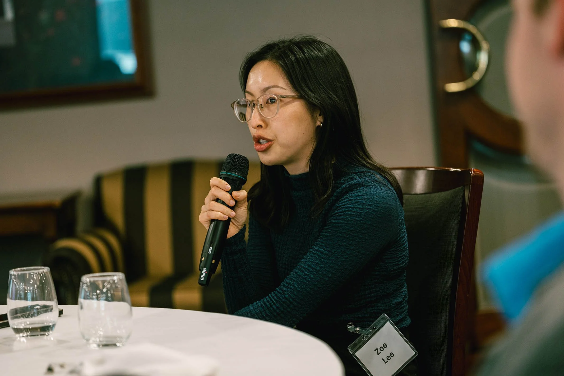 A woman with black hair and glasses speaking into a microphone during a panel or discussion, seated at a table with three glasses of water, wearing a dark sweater, and a name tag that reads "Zoe Lee."