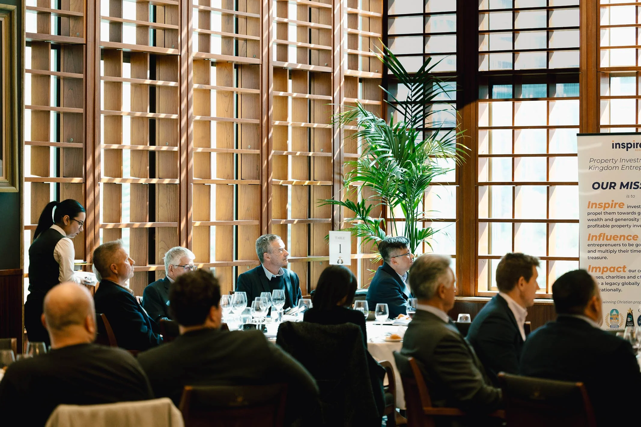 Business meeting or conference inside a well-lit room with large wooden windows, a tall green plant, and a partially visible poster on the right side.
