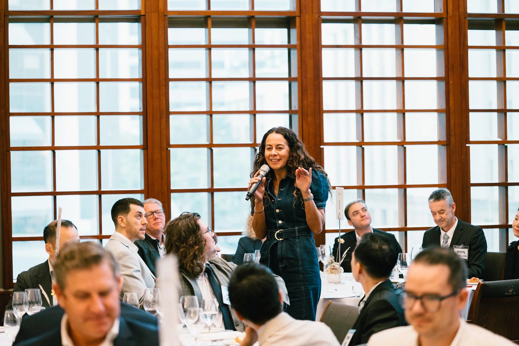 A woman with long curly hair speaks into a microphone at a formal event, surrounded by seated attendees in a room with large wooden windows.